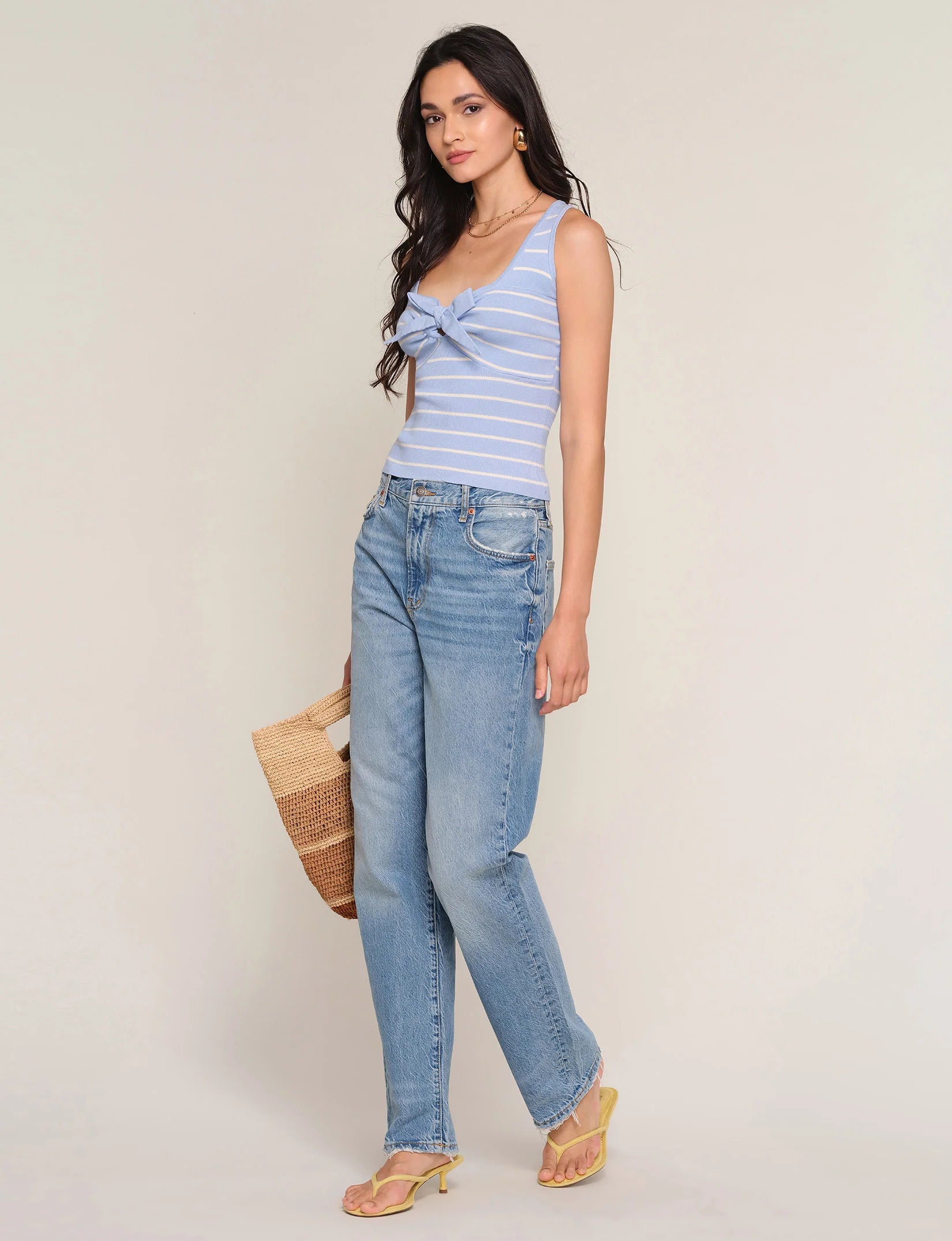 A woman stands against a light background wearing the Heartloom Tiffy Top—a slim fit, light blue and white striped tank with a front bow—paired with light blue jeans, yellow heeled sandals, and holding a woven tote bag.