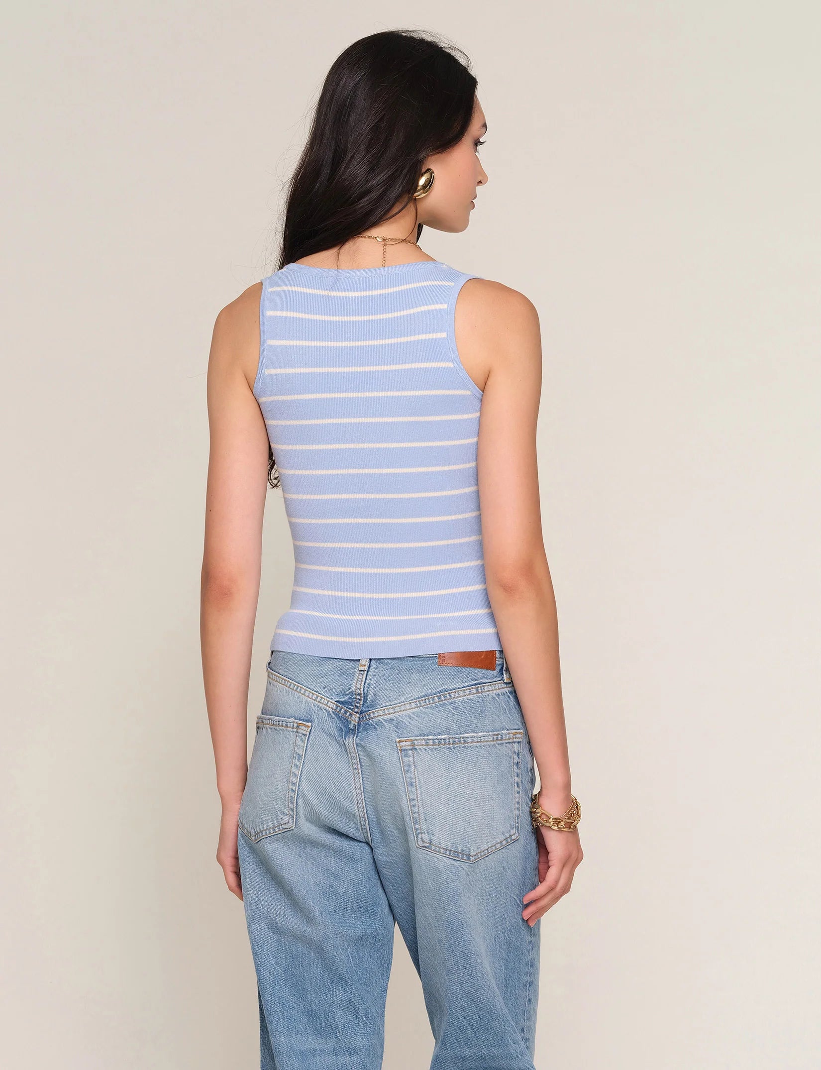 A woman with long dark hair models the Heartloom Tiffy Top—a slim fit, light blue sleeveless top with white stripes—paired with light blue jeans. She accessorizes with gold earrings and a bracelet against a plain, light background.