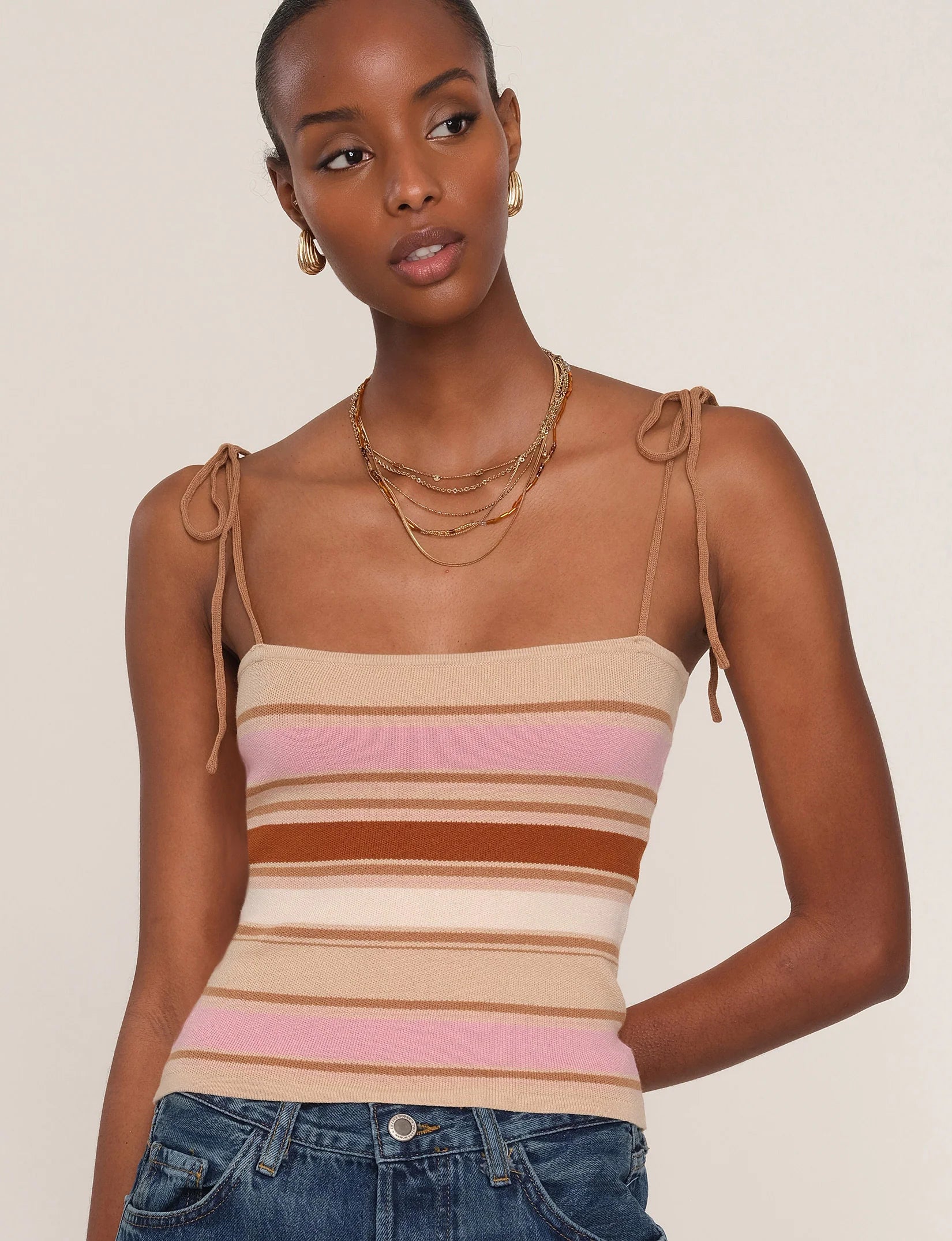 A woman wearing gold hoop earrings, layered necklaces, and the Heartloom Teyana Top in Pink Multi pairs it with blue jeans, standing against a light background.