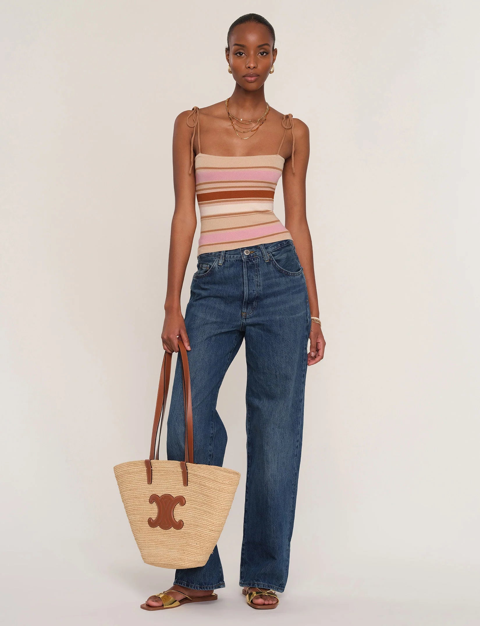 A woman stands against a light background wearing the Heartloom Teyana Top—a pink and beige yarn-dye stripe tank—paired with blue wide-leg jeans, tan sandals, a straw tote bag with brown handles and logo, plus layered necklaces.