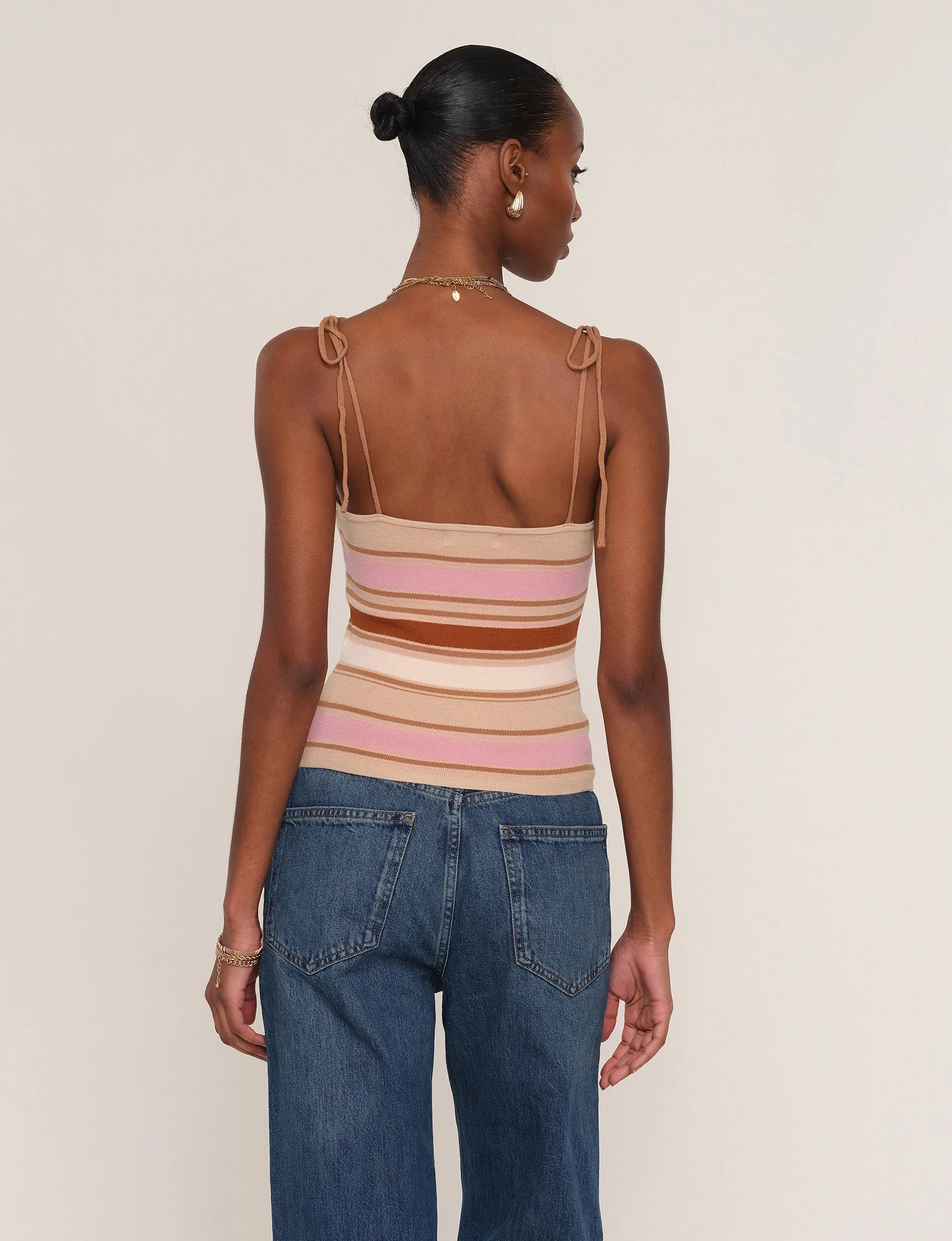 A woman faces away, wearing the Heartloom Teyana Top—a slim fit, striped knit with tied shoulder straps—paired with blue jeans. Her hair is in a neat bun, and she accessorizes with gold hoop earrings, a necklace, and a bracelet.