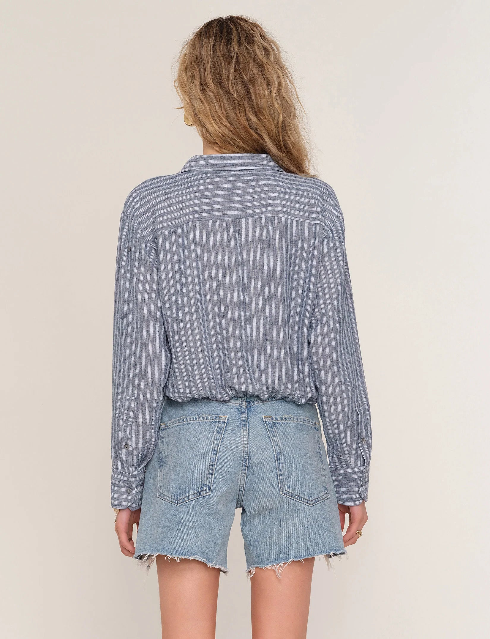 A person with wavy blonde hair is shown from behind, wearing the Heartloom Rene Shirt—a blue and white striped button-down—tucked into high-waisted, light-wash denim shorts with frayed hems.