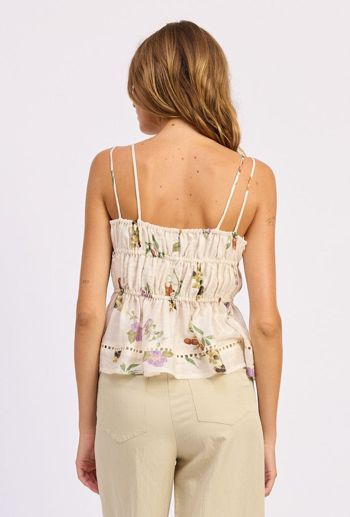 A woman with long, wavy hair models the En Saison Tusla Peplum Top—sleeveless with a floral print and adjustable tie-shoulder straps—paired with cream pants, standing with her back to the camera against a plain light background.