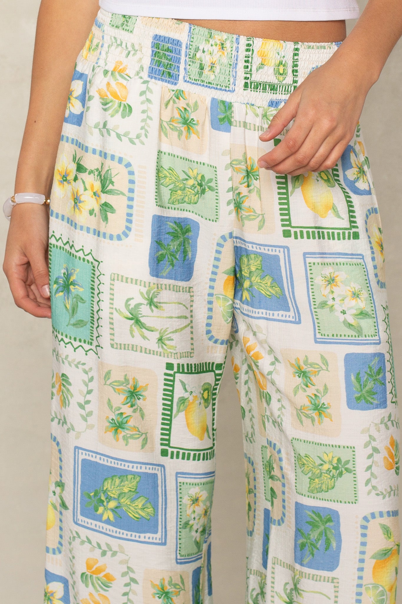 A person wears Isla's Hattie Beach Pant—loose, high-waisted, full-length with a tropical patchwork of green leaves, yellow flowers, and blue-green squares. A hand rests on its smocked waistband, highlighting the straight-leg style.