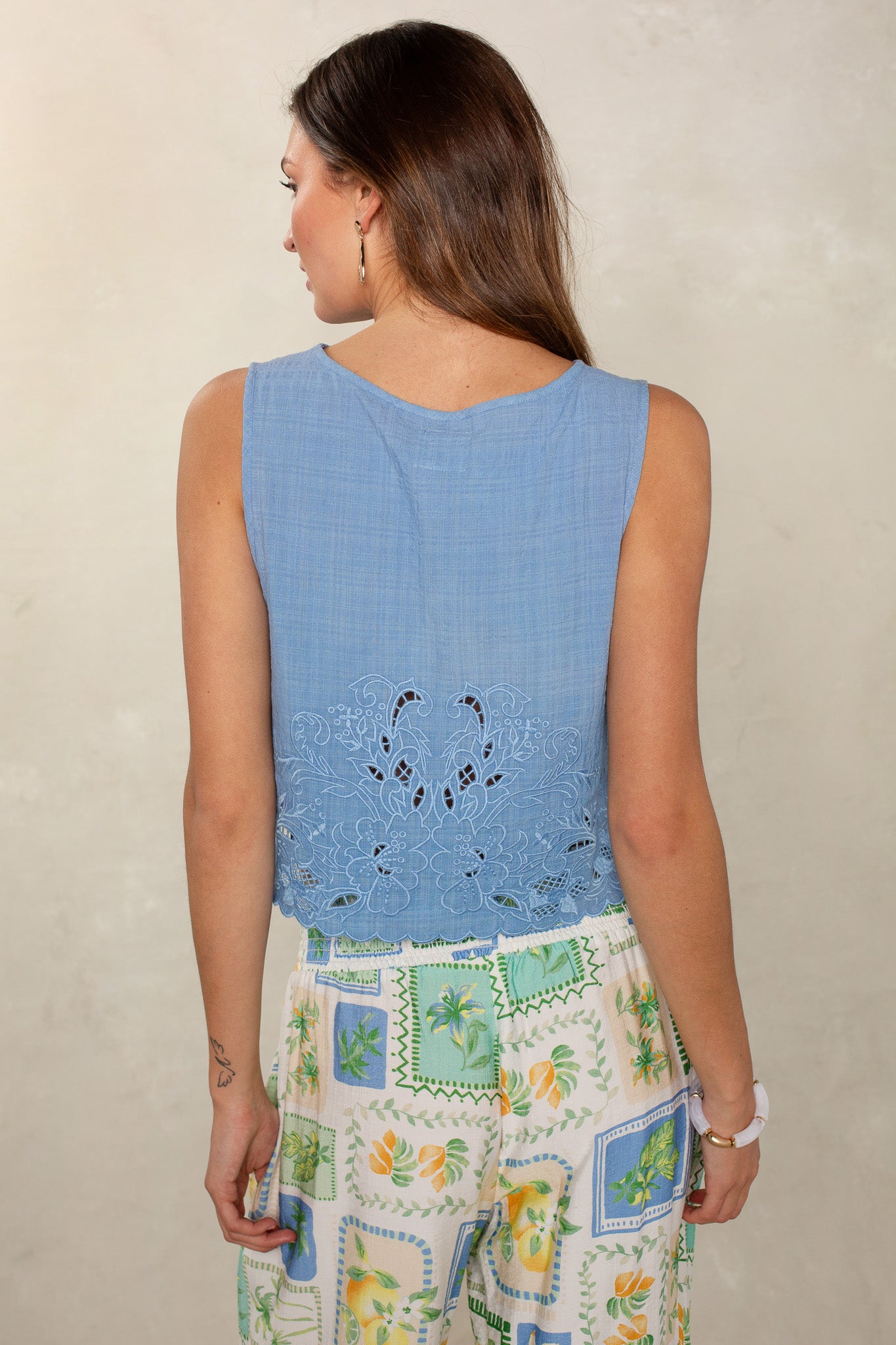A woman faces away from the camera, wearing Isla’s sleeveless Desert Sunset Top with lace cutout details on the back and high-waisted pants with a colorful botanical patchwork print for a sophisticated, relaxed look.