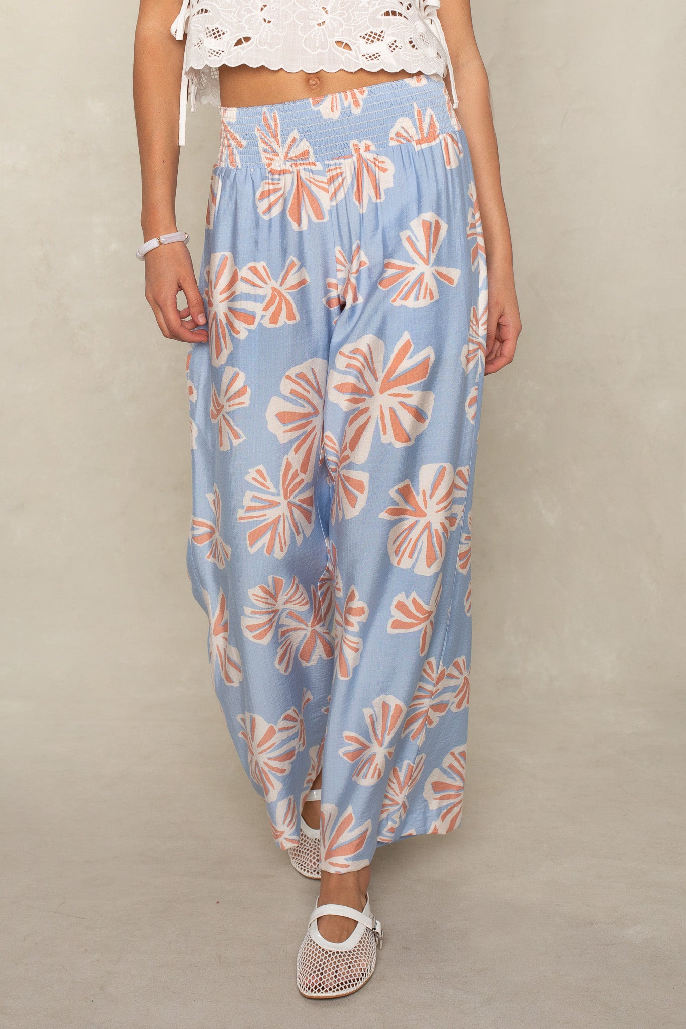 A person in Isla's Hattie Beach Pant—loose, light blue straight-leg pants with a pink and white floral pattern and smocked waistband—plus a white lace crop top and white mesh shoes stands against a neutral background.