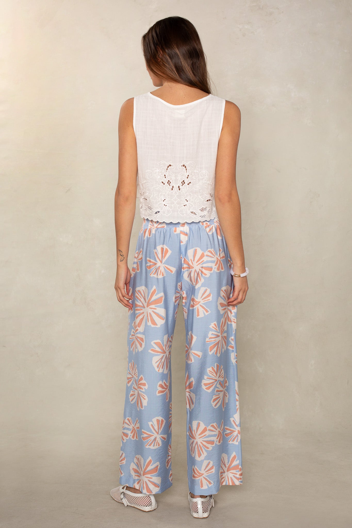 A woman stands facing away, wearing a sleeveless white lace-detailed top and Isla Hattie Beach Pant—loose, full-length pants with large pink and white floral patterns—paired with white shoes.
