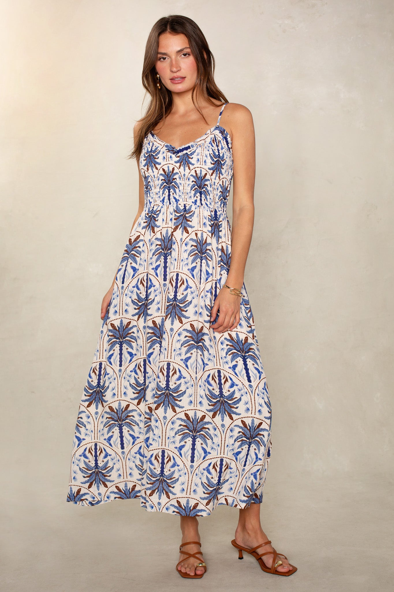 A woman wears the Isla Lana Smocked Easy Midi Dress, sleeveless and ankle-length with blue and brown palm tree patterns. She pairs it with brown strappy sandals; her long wavy brown hair contrasts a plain light background.