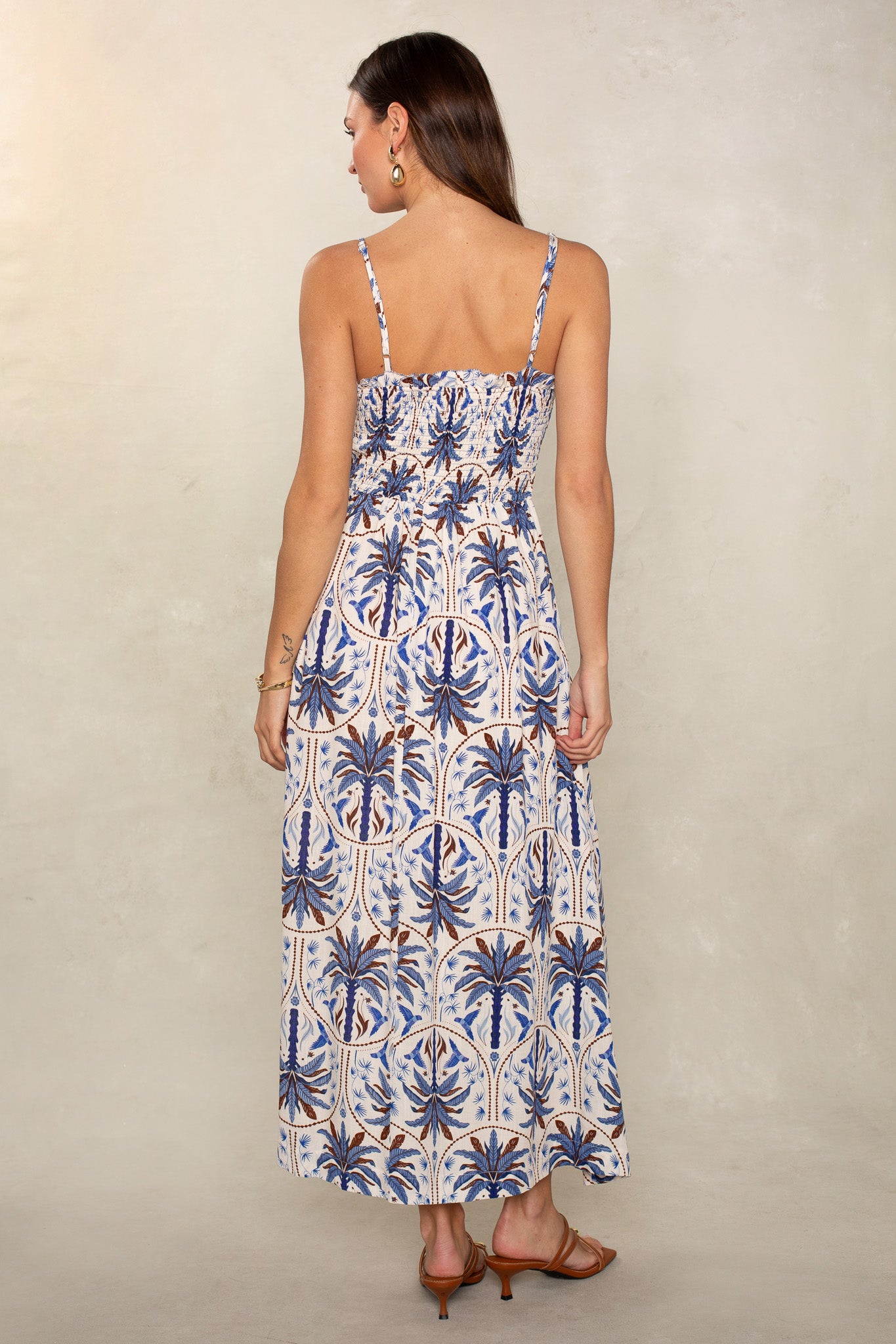 A woman with long brown hair, wearing Isla’s Lana Smocked Easy Midi Dress in a blue and brown palm tree pattern, stands with her back to the camera on a neutral background. She pairs the sleeveless midi dress with brown heeled sandals.