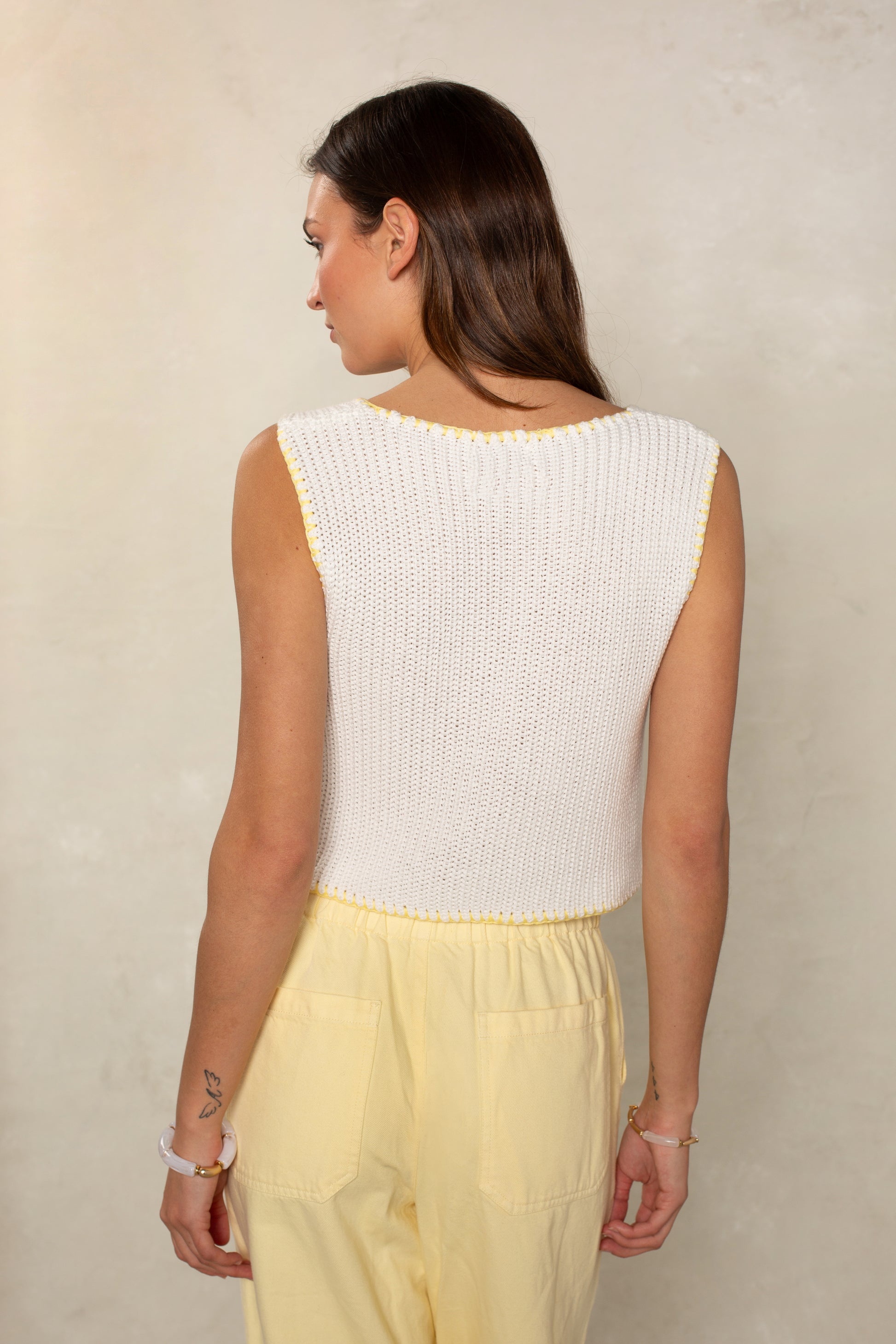 A woman with long brown hair stands with her back to the camera, wearing Isla’s Contrast Sweater Tank—a sleeveless white knit top with a cropped fit—and yellow pants with patch pockets against a light, neutral background.