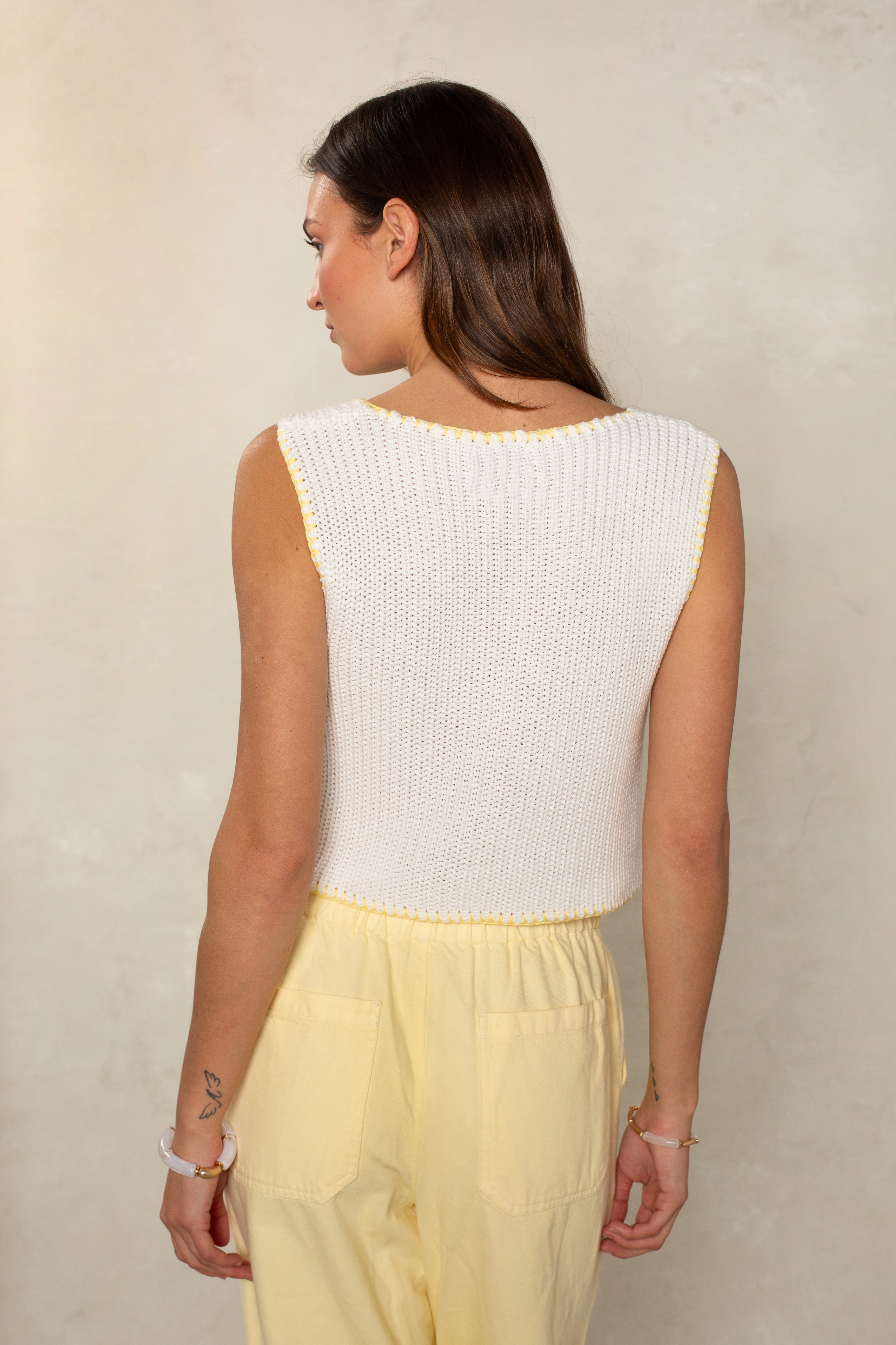 A woman with long brown hair stands with her back to the camera, wearing Isla’s Contrast Sweater Tank—a sleeveless white knit top with a cropped fit—and yellow pants with patch pockets against a light, neutral background.