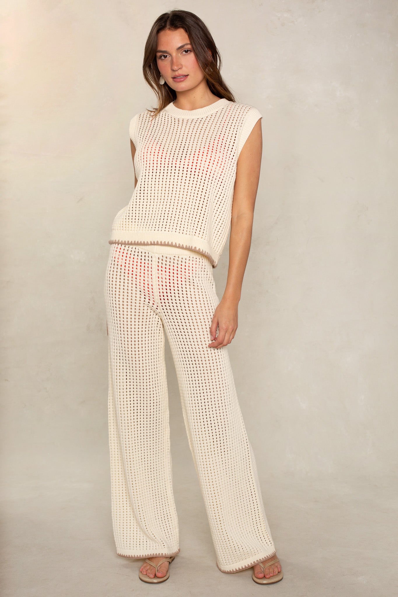 A woman with long brown hair stands against a light textured background, wearing Isla's Eileen Cover Up Sweater—a sleeveless, cream crochet top—paired with matching wide-leg pants and sandals for a relaxed, versatile look.