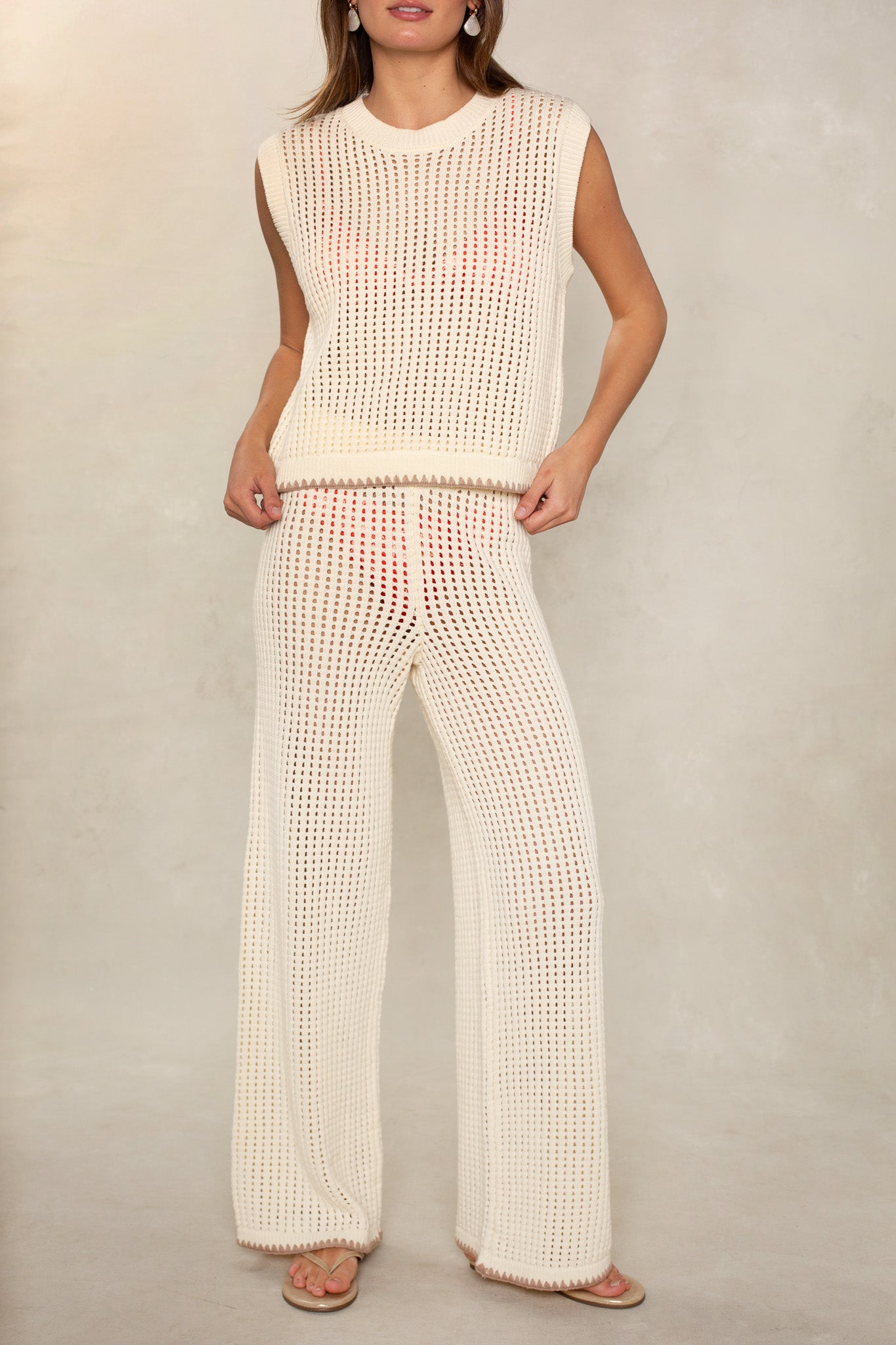 A woman stands relaxed against a neutral background, wearing the Isla Eileen Cover Up Sweater—a sleeveless cream crochet top styled over a visible pink bikini, paired with matching wide-leg pants. Perfect as a swimsuit cover up.