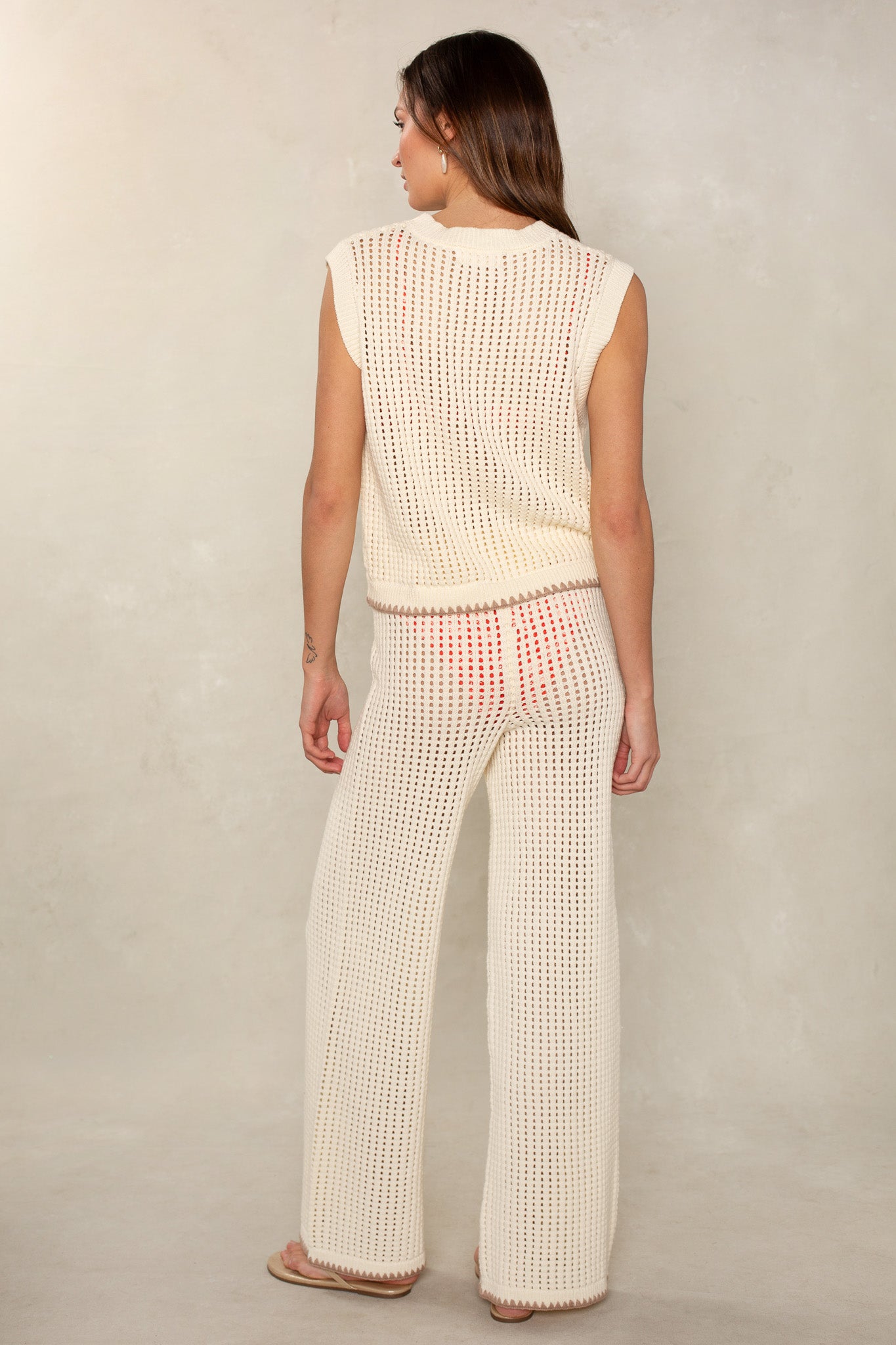 A woman is seen from behind wearing the Isla Eileen Crochet Coverup Pant, a cream open-knit style that reveals a hint of red underwear beneath.