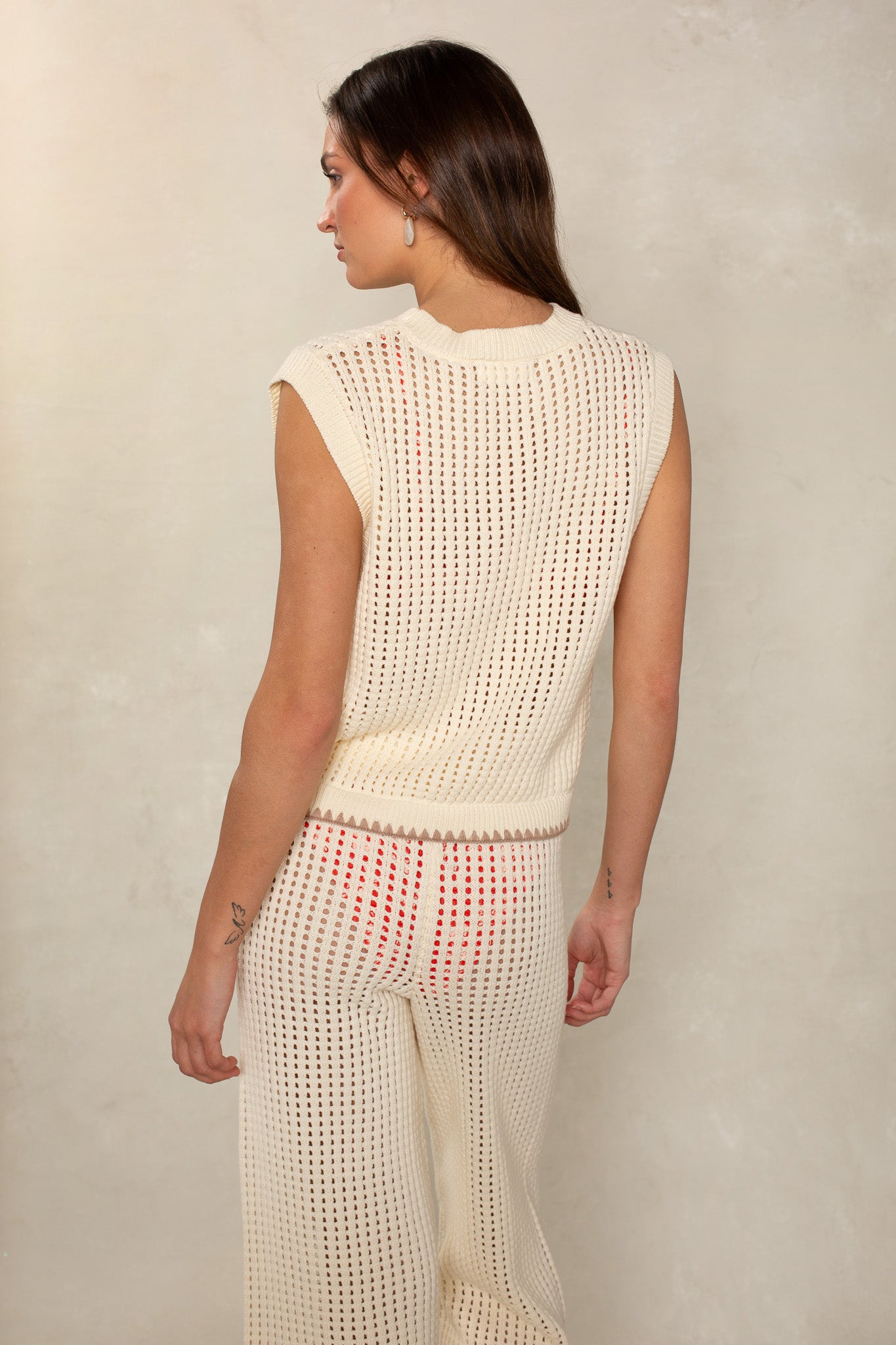 A woman faces away, wearing the Isla Eileen Cover Up Sweater—a sleeveless, cream crochet top that's lightweight and slightly sheer—paired with matching pants and red undergarments, set against a light beige wall.