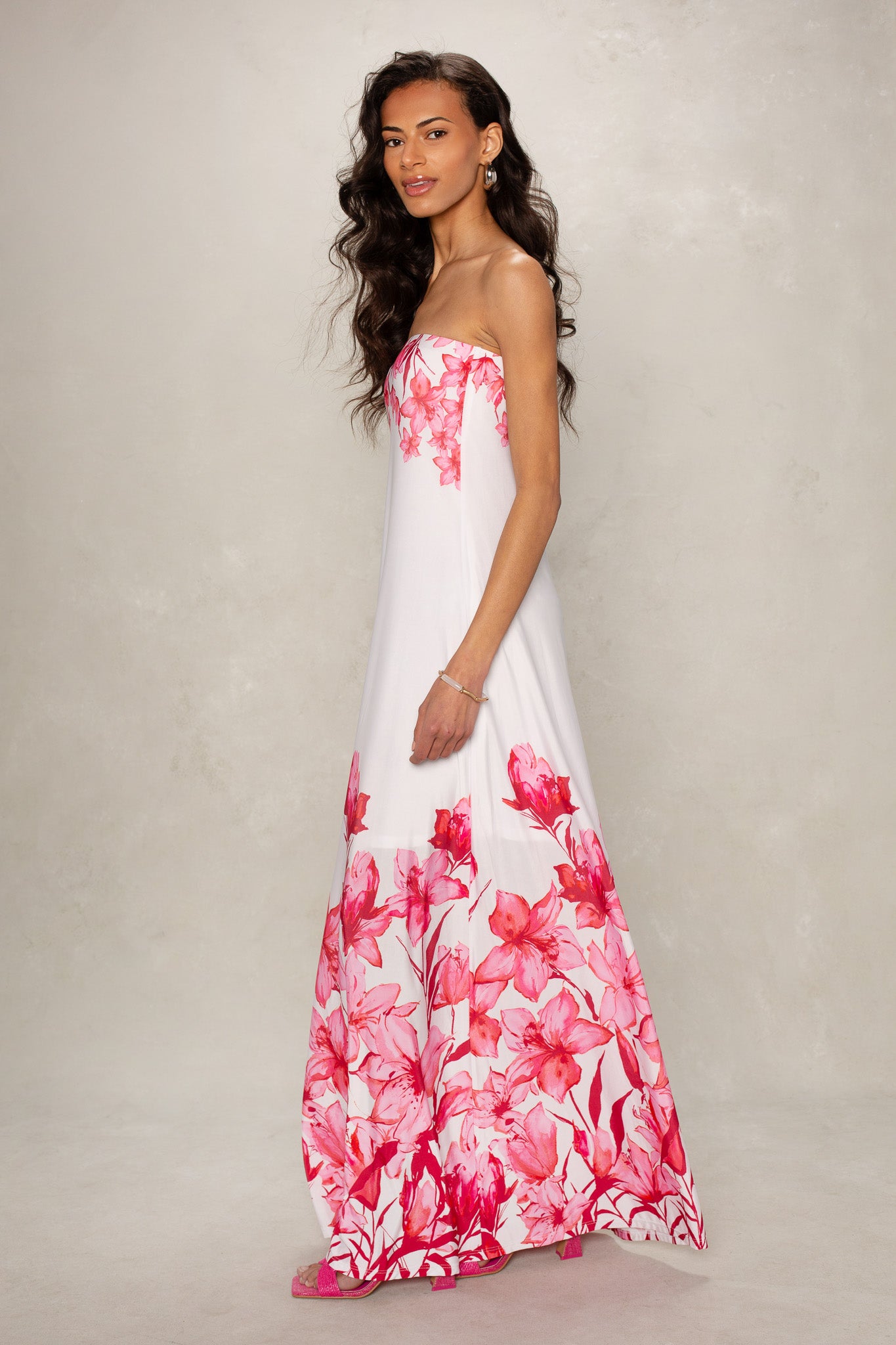 A woman with long wavy hair wears the Isla Flora Tube Maxi Dress, a strapless white maxi adorned with pink floral patterns on the hem and bodice, as she stands against a light neutral background.