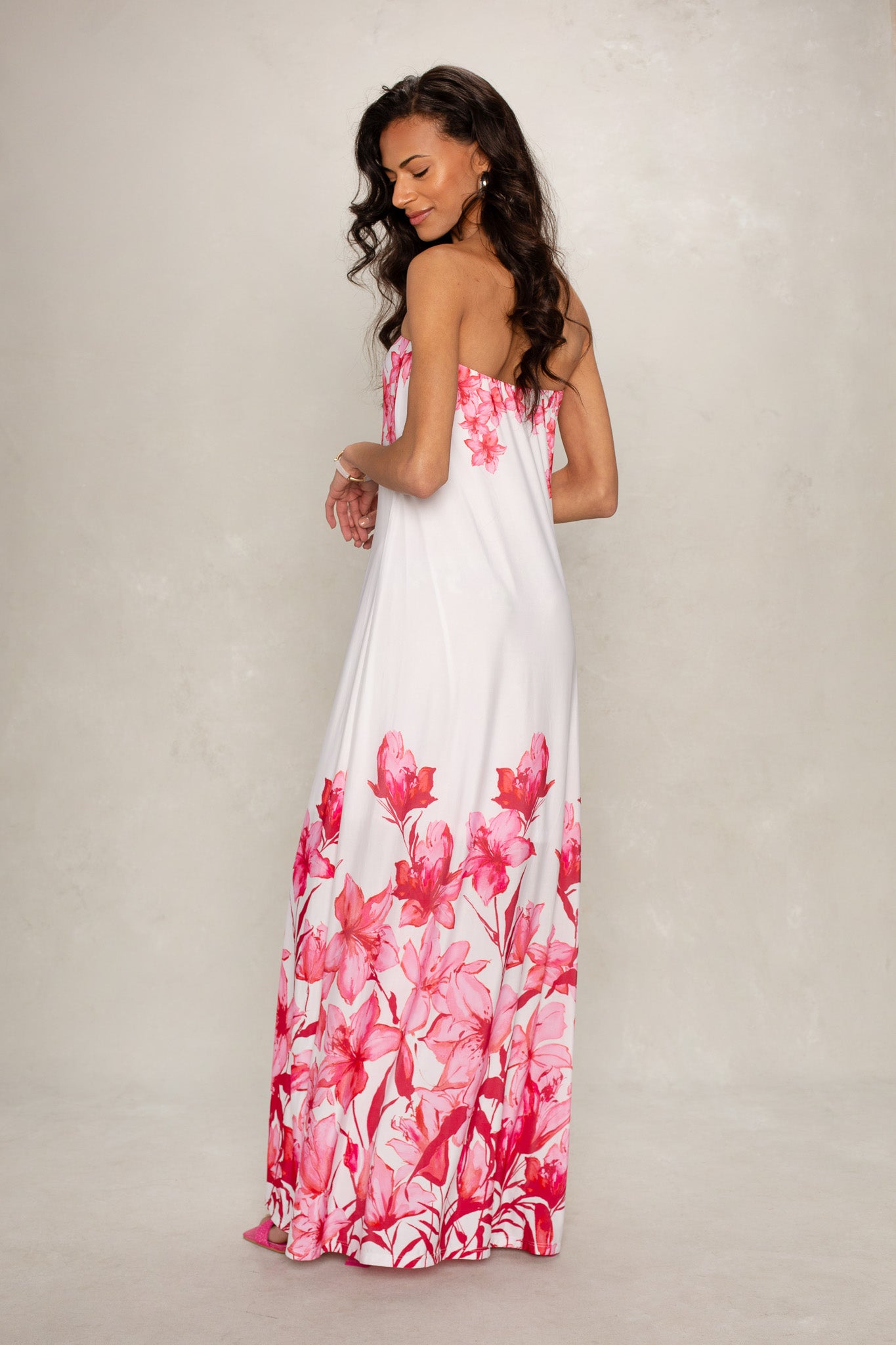 A woman with long dark hair models the Isla Flora Tube Maxi Dress, a strapless maxi featuring pink floral patterns along the bottom and back, as she stands against a neutral background, looking over her shoulder with a gentle smile.