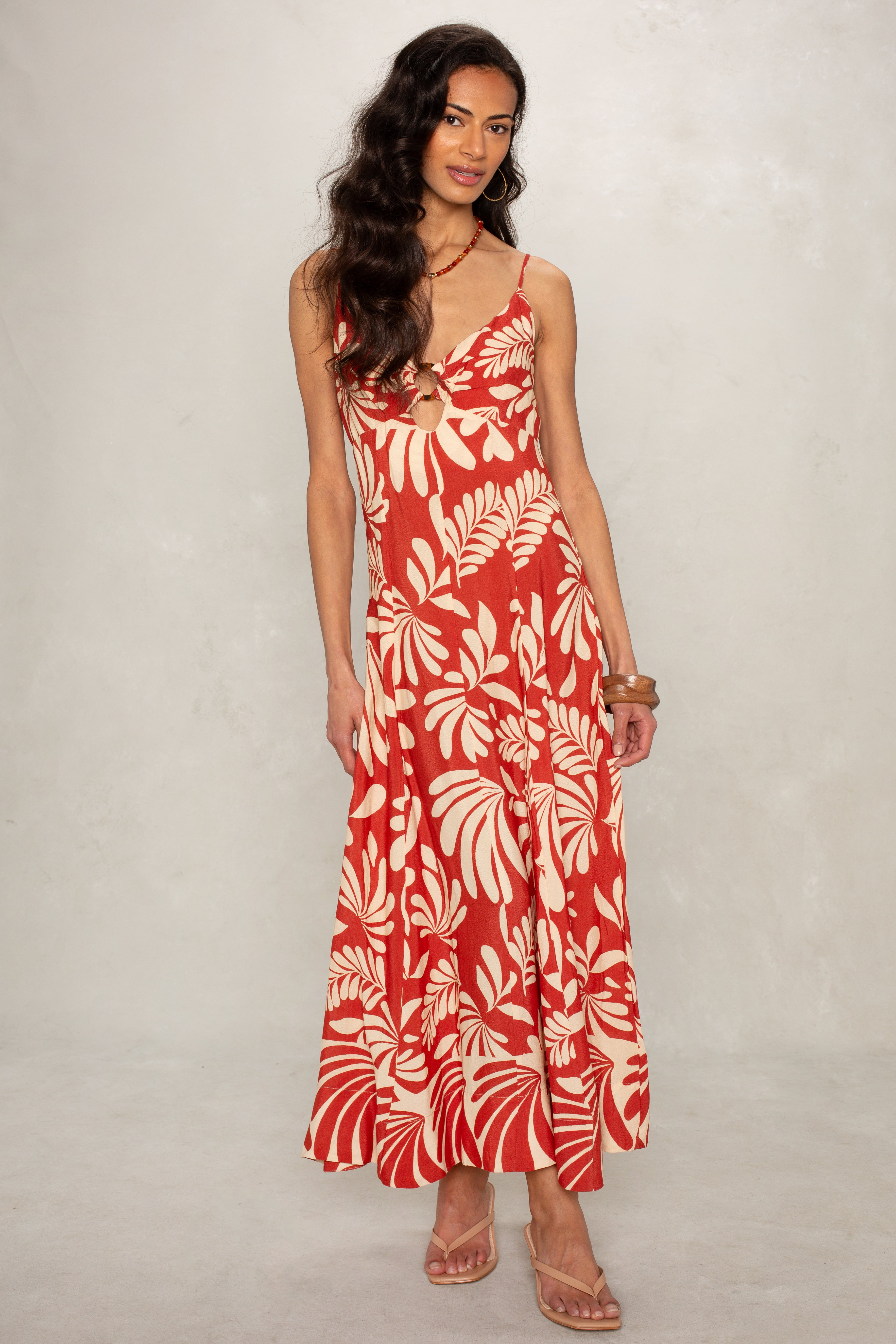 A woman stands against a light background wearing the Isla Ariel O-ring Midi Dress in red and white floral print with spaghetti straps, paired with beige sandals and loose wavy hair.