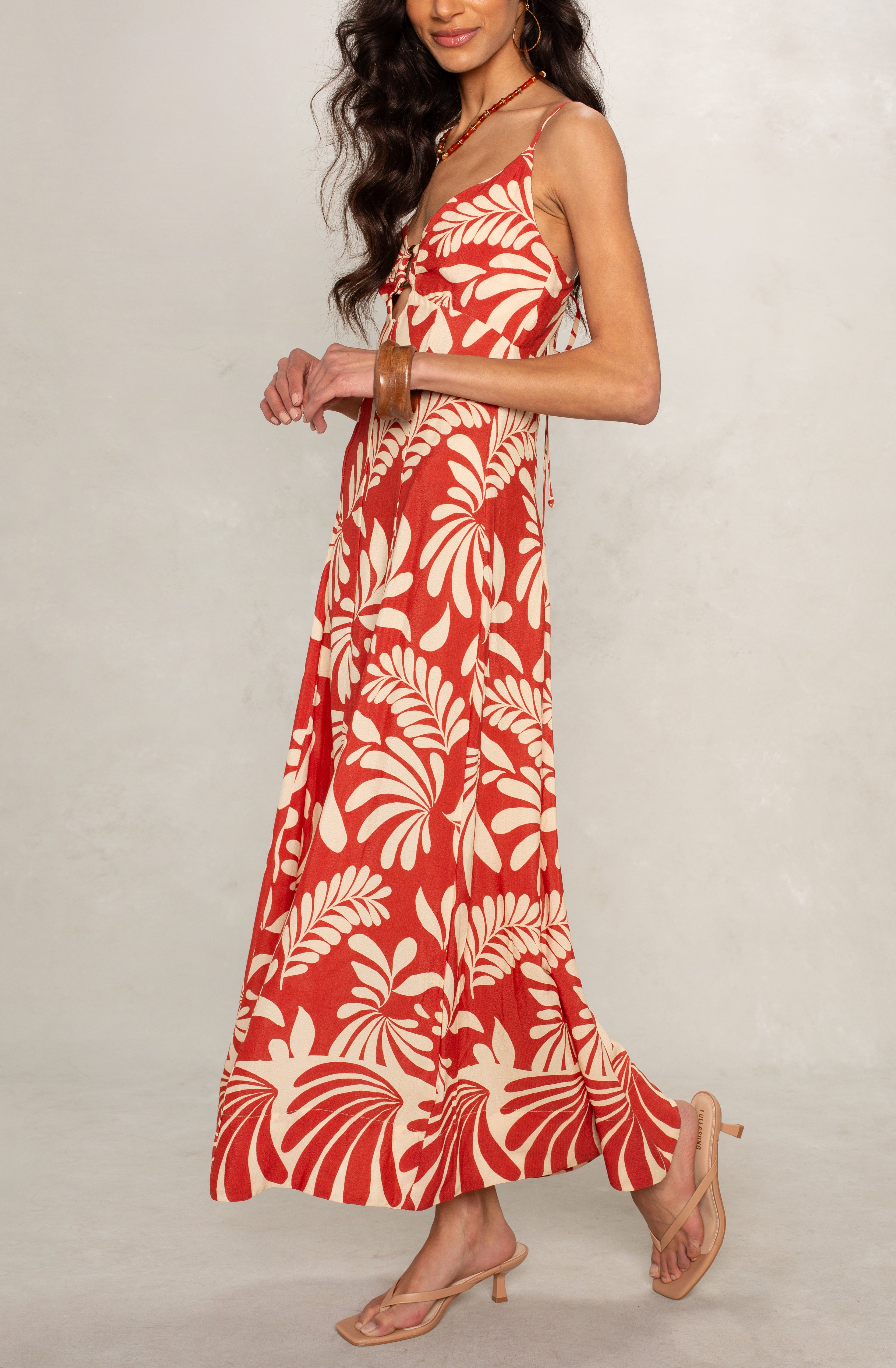 A woman models the Isla Ariel O-ring Midi Dress, featuring a red and beige tropical pattern with thin straps, styled with bracelets and nude heeled sandals against a light neutral background.