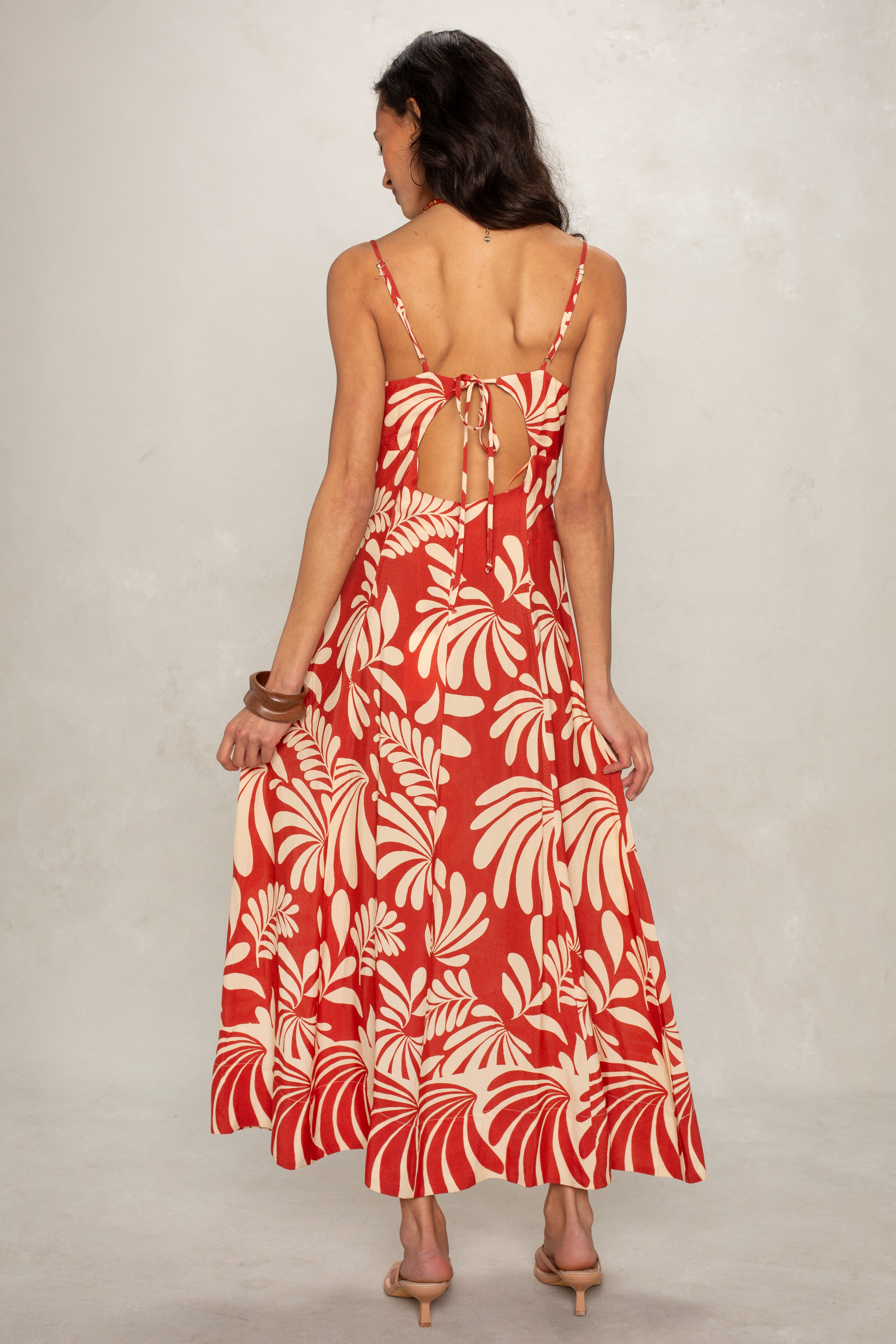 A woman with wavy brown hair wears the Isla Ariel O-ring Midi Dress—a red and beige sleeveless dress featuring a bold leaf pattern, open back with string ties, and sandals—standing against a plain background.