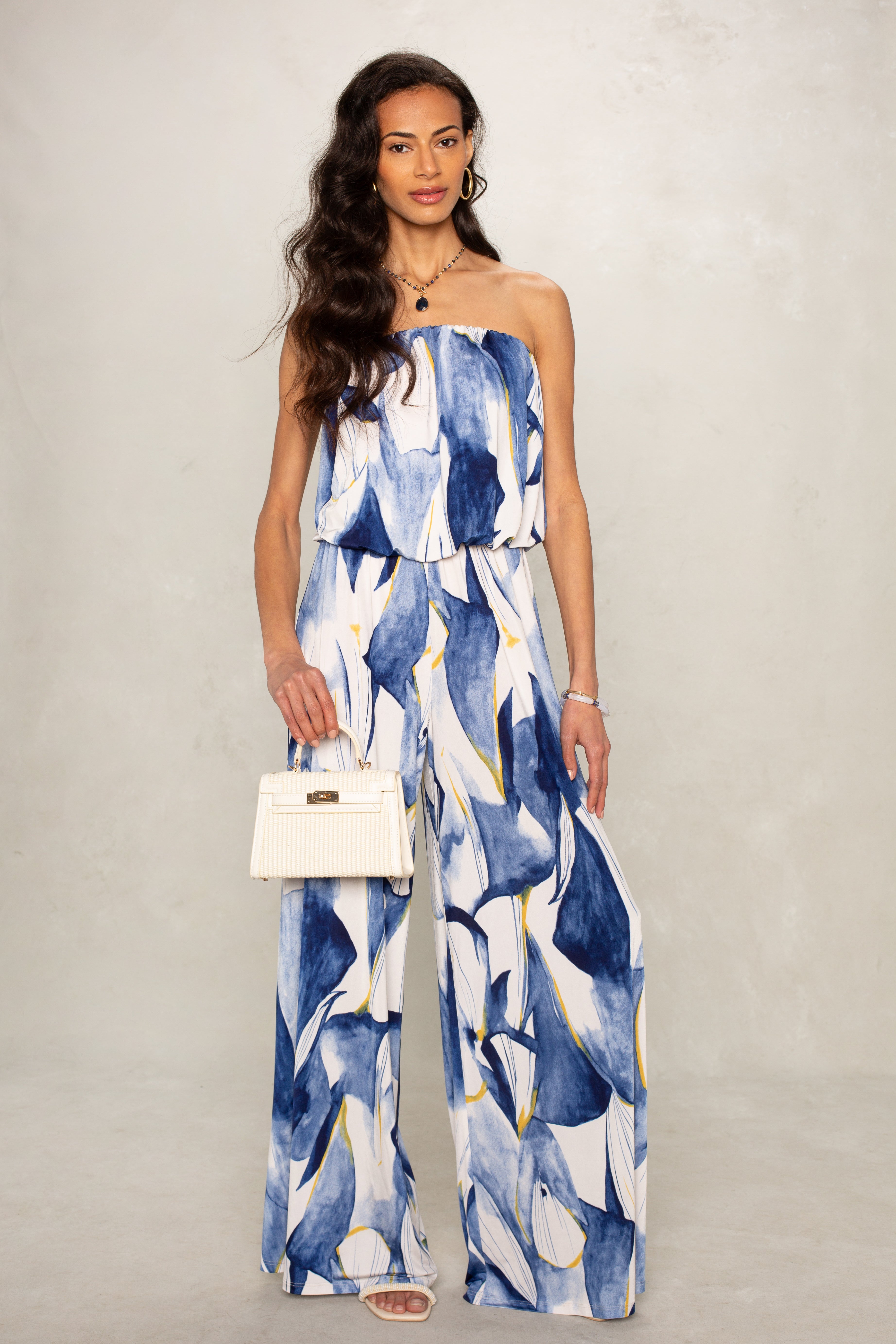 A woman wears the Isla Ari Strapless Easy Jumpsuit—a blue and white modern essential—paired with white sandals, a small white handbag, layered necklaces, and bracelets. Her long wavy hair completes the look against a light background.