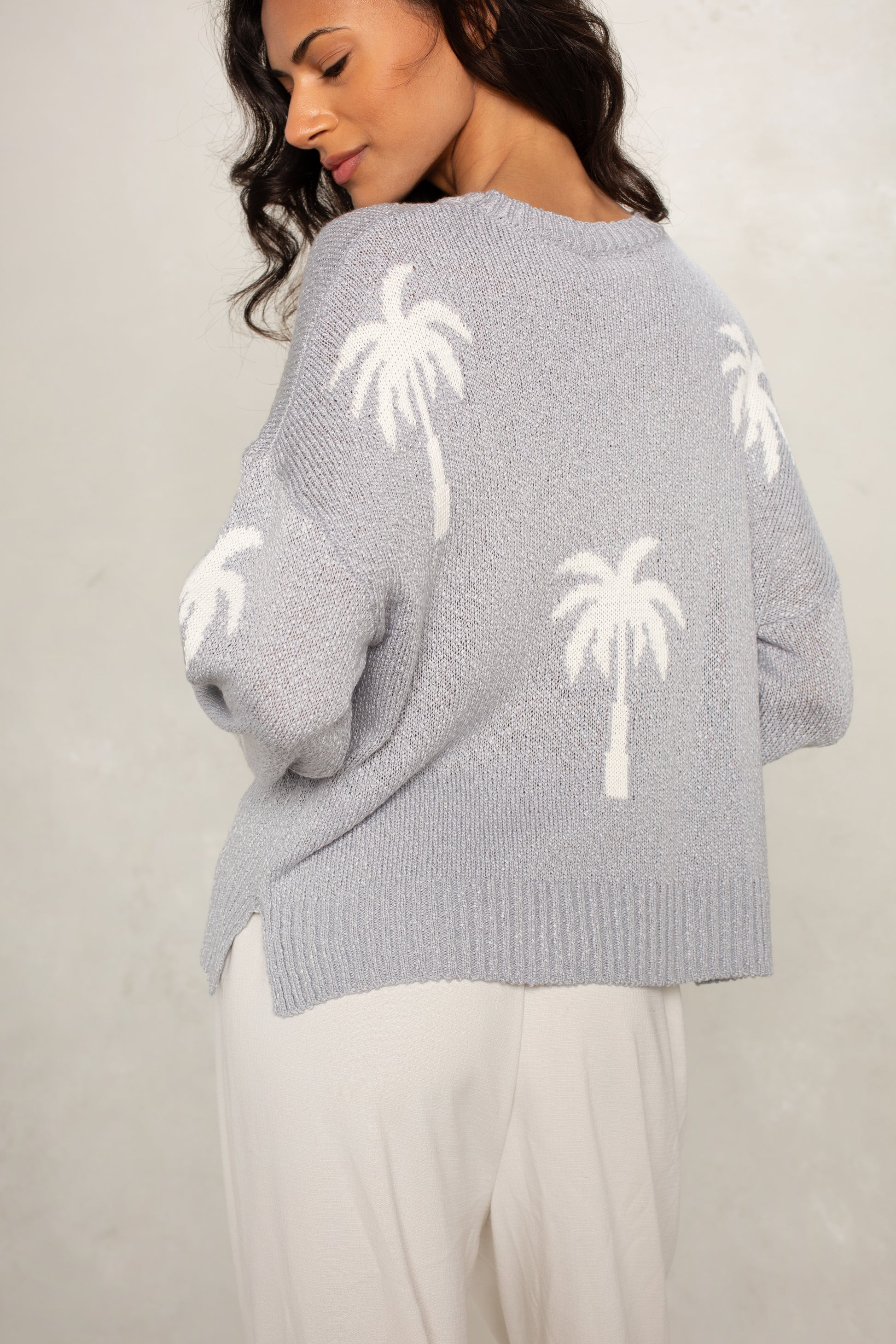 A woman with long dark hair models the Isla Palm Beaches Sweater, a loose light gray top with white palm tree designs, paired with light-colored pants, against a neutral backdrop.