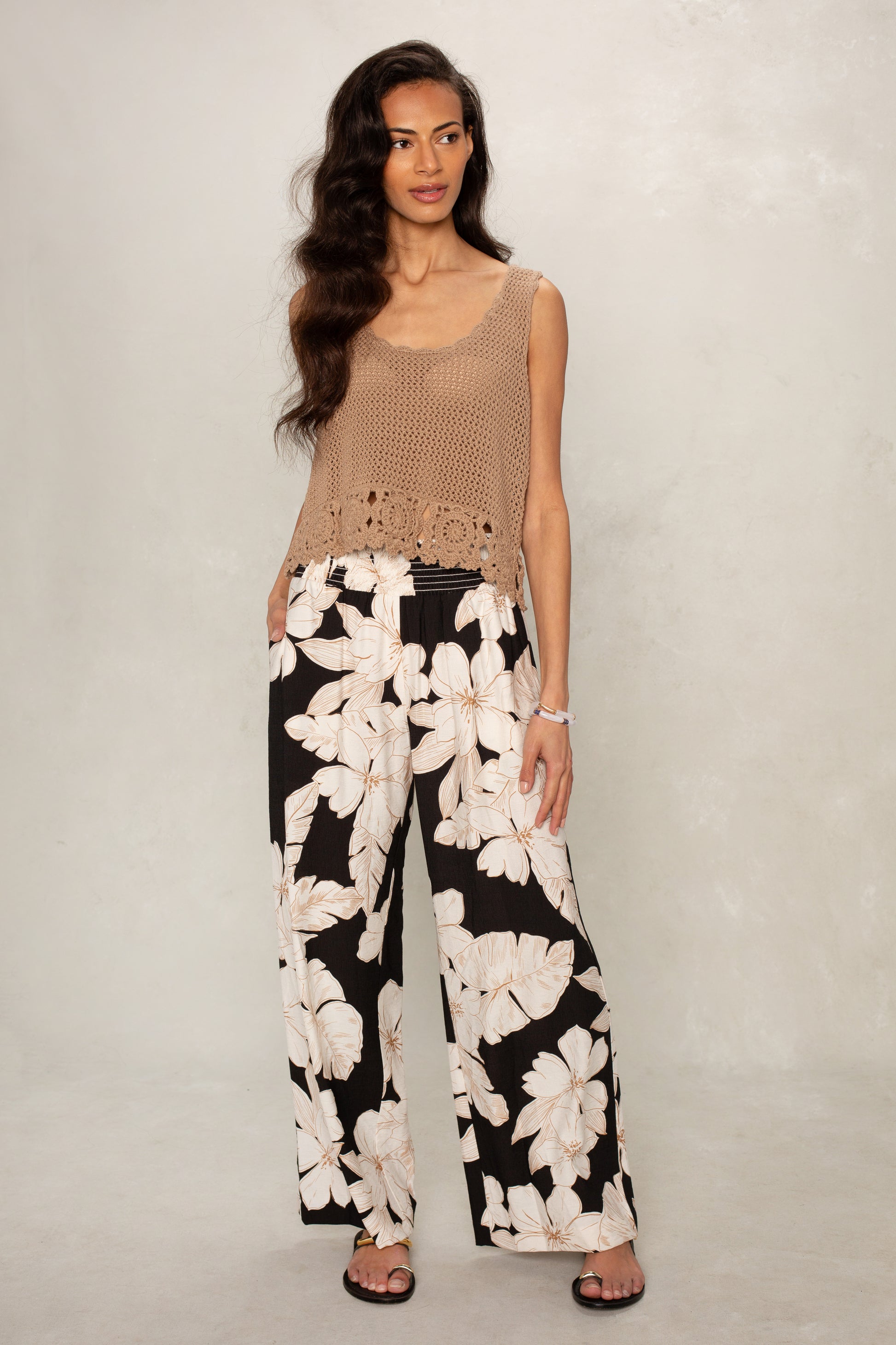 A woman with long, wavy hair stands against a light background wearing Isla’s Hattie Beach Pant in Black Floral with a sleeveless beige crochet top and black sandals.