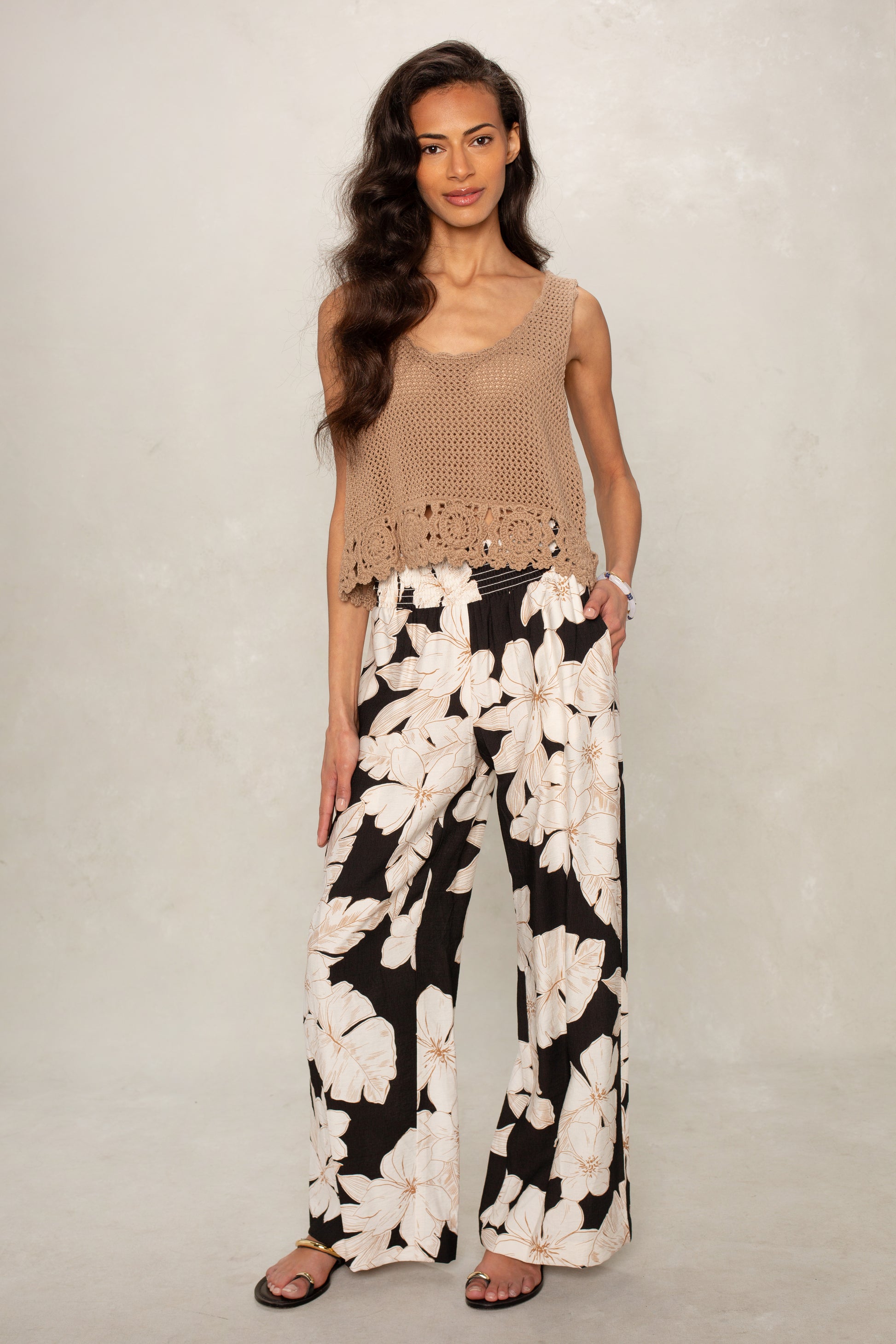A woman with long wavy hair stands against a light background wearing Isla's Hattie Beach Pant—a loose, full-length pant with a bold white floral print and comfy smocked waistband—paired with sandals and a sleeveless brown crochet top.