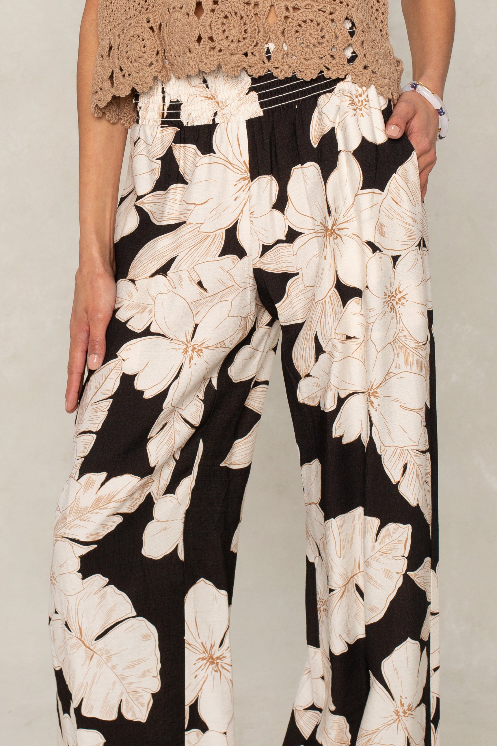 Wearing Isla's Hattie Beach Pant with a large white floral print and smocked waistband, paired with a brown crochet top, they stand against a light background—one hand in their pocket, the other by their side.