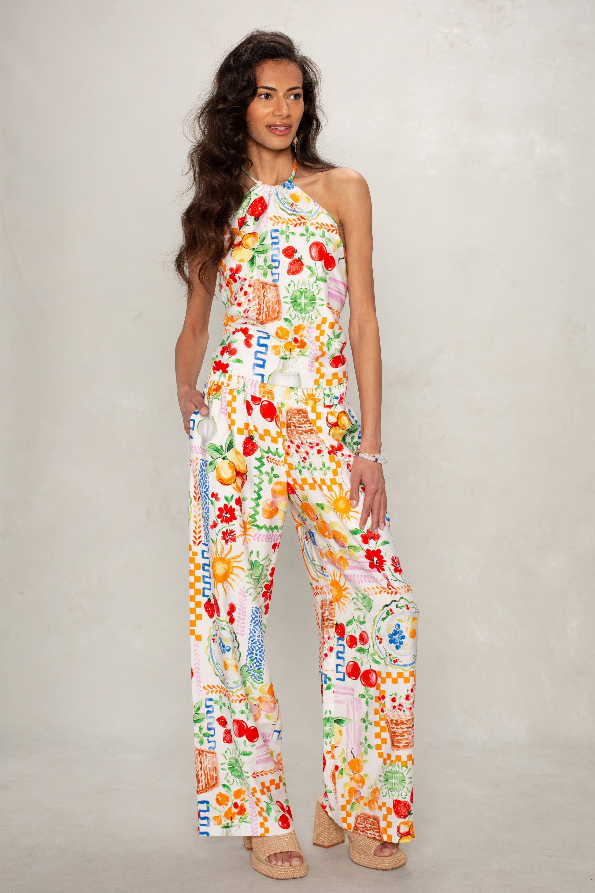 A woman poses against a light gray background wearing Isla’s Hattie Beach Pant in Multi—a colorful, straight-leg, full-length pant with a fruit and floral print and smocked waistband. She smiles slightly with one hand in her pocket.