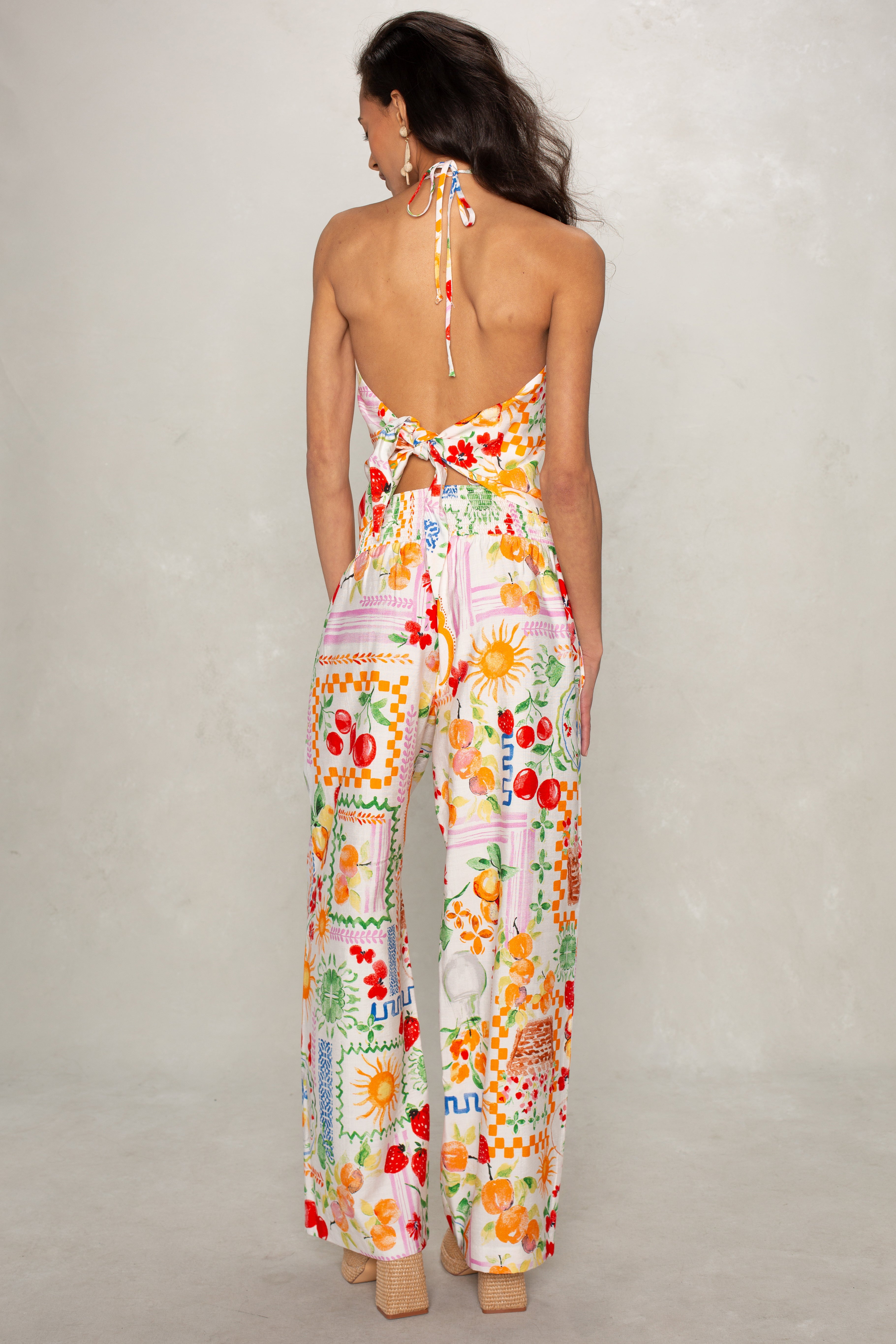A woman with long dark hair wears Isla’s Hattie Beach Pant, featuring bright floral and geometric patterns, a smocked waistband, and full-length cut. She faces away in sandals, showcasing the colorful design.
