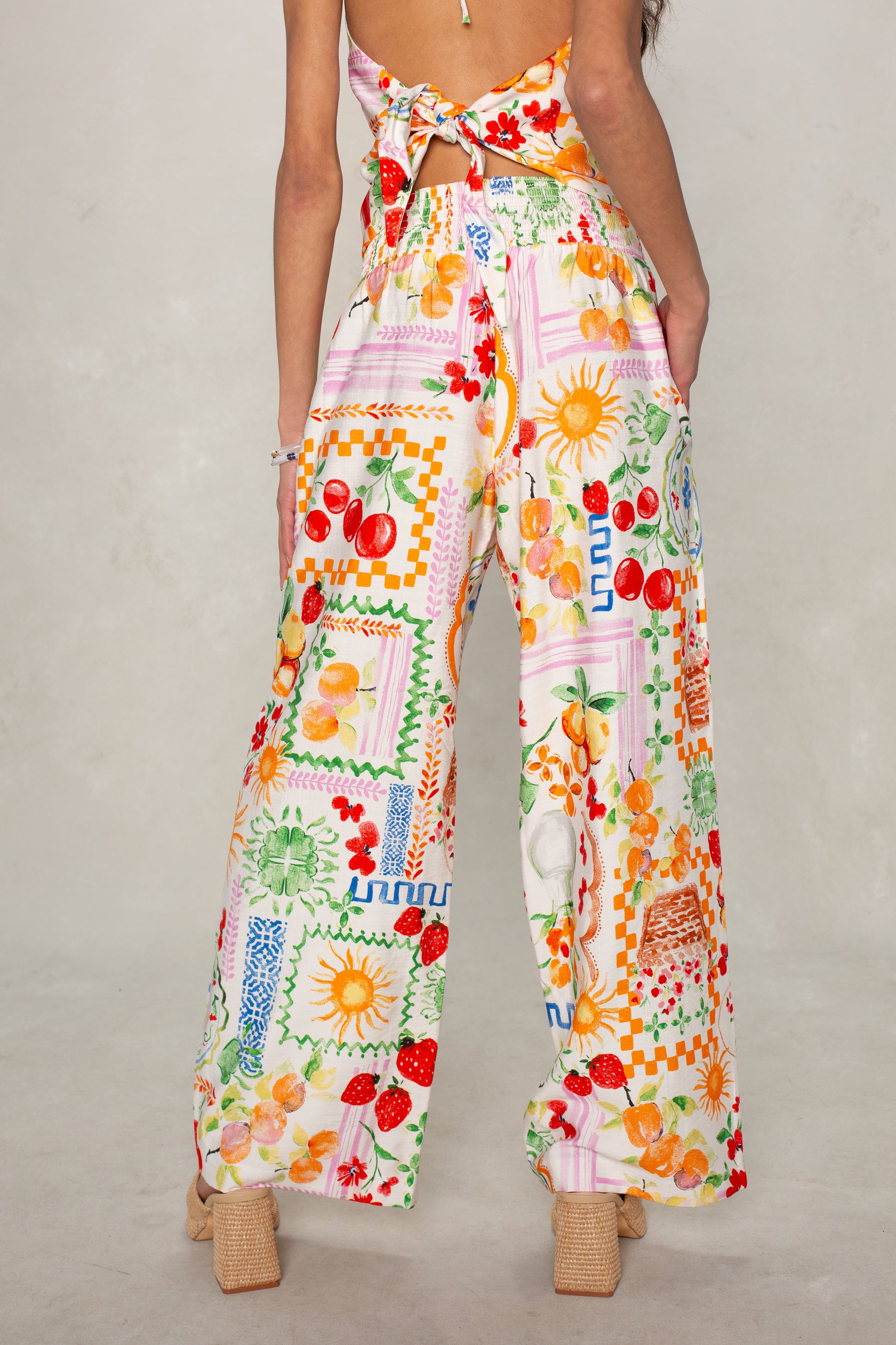 A woman faces away wearing Isla's Hattie Beach Pant, featuring a smocked waistband and colorful fruit and floral patchwork print. She pairs them with beige heeled sandals and a back-tied sleeveless top.
