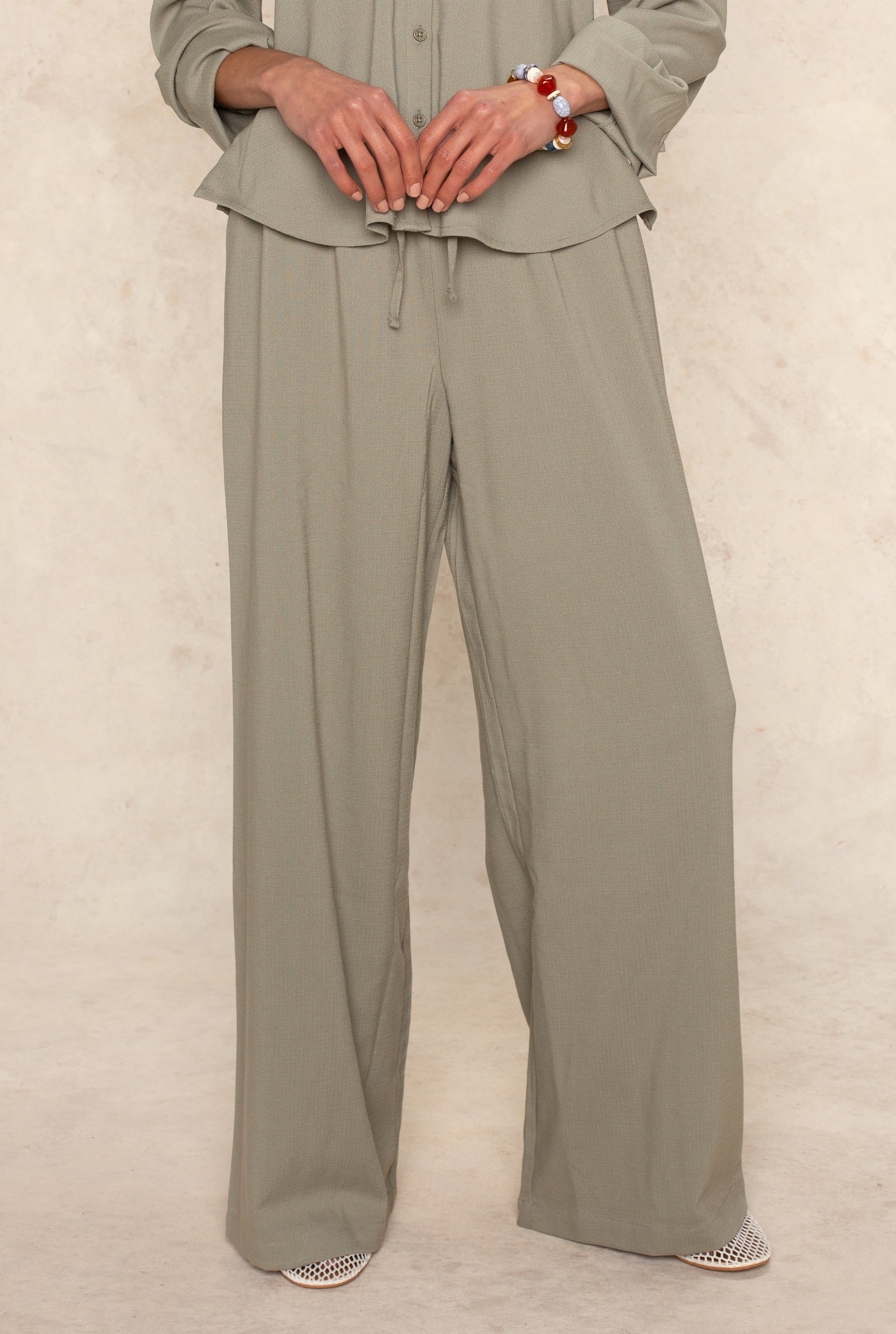 A person in Georgie’s sage green Seaside Pant stands against a light beige background—an airy, breathable look ideal for warm-weather outings. Their loosely clasped hands and only the lower half of their body are visible.