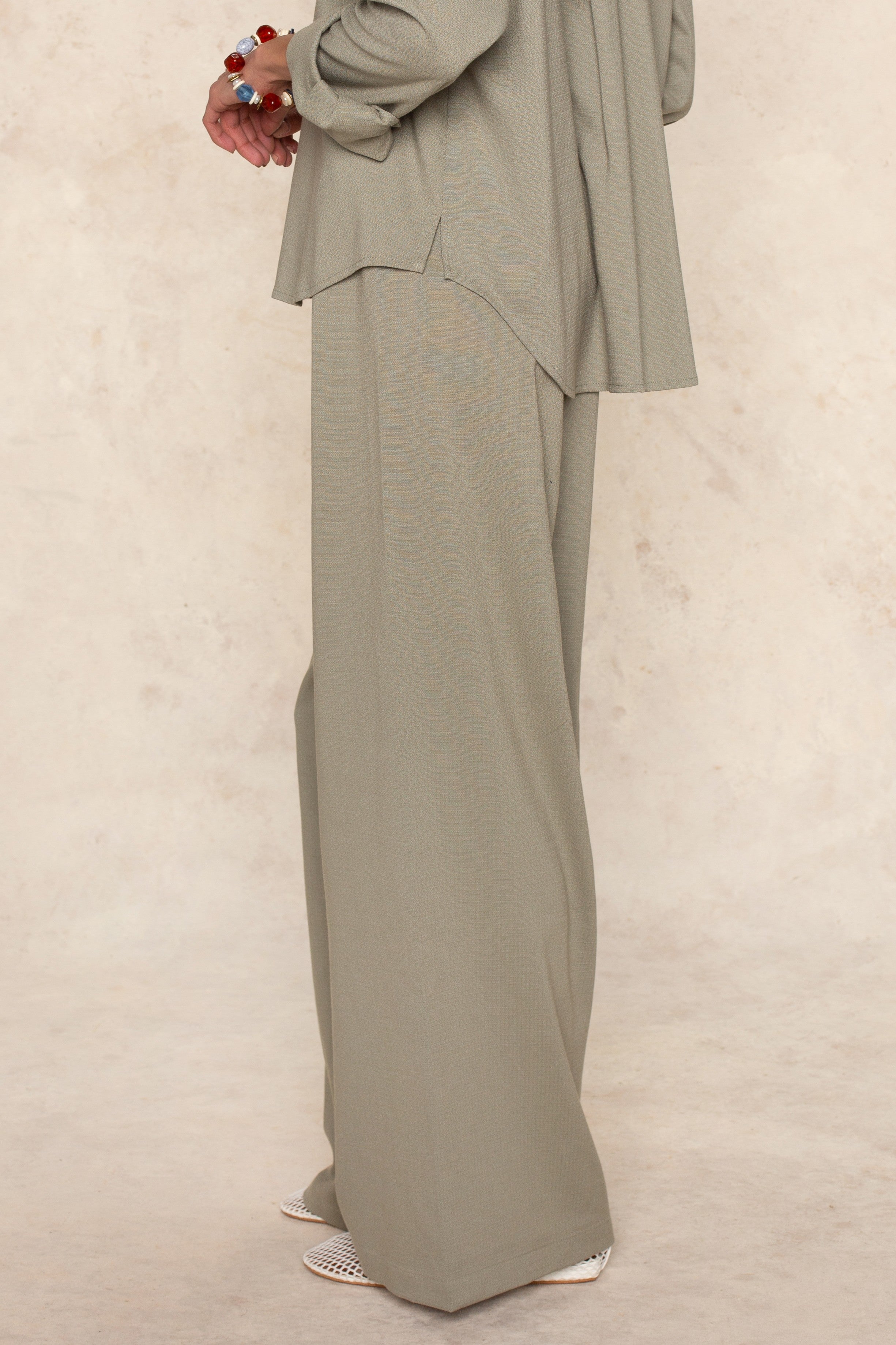 Wearing the Georgie Seaside Pant and a matching light olive green top, paired with white pointed-toe shoes and a chunky bracelet—an ideal warm-weather casual look set against a neutral backdrop.