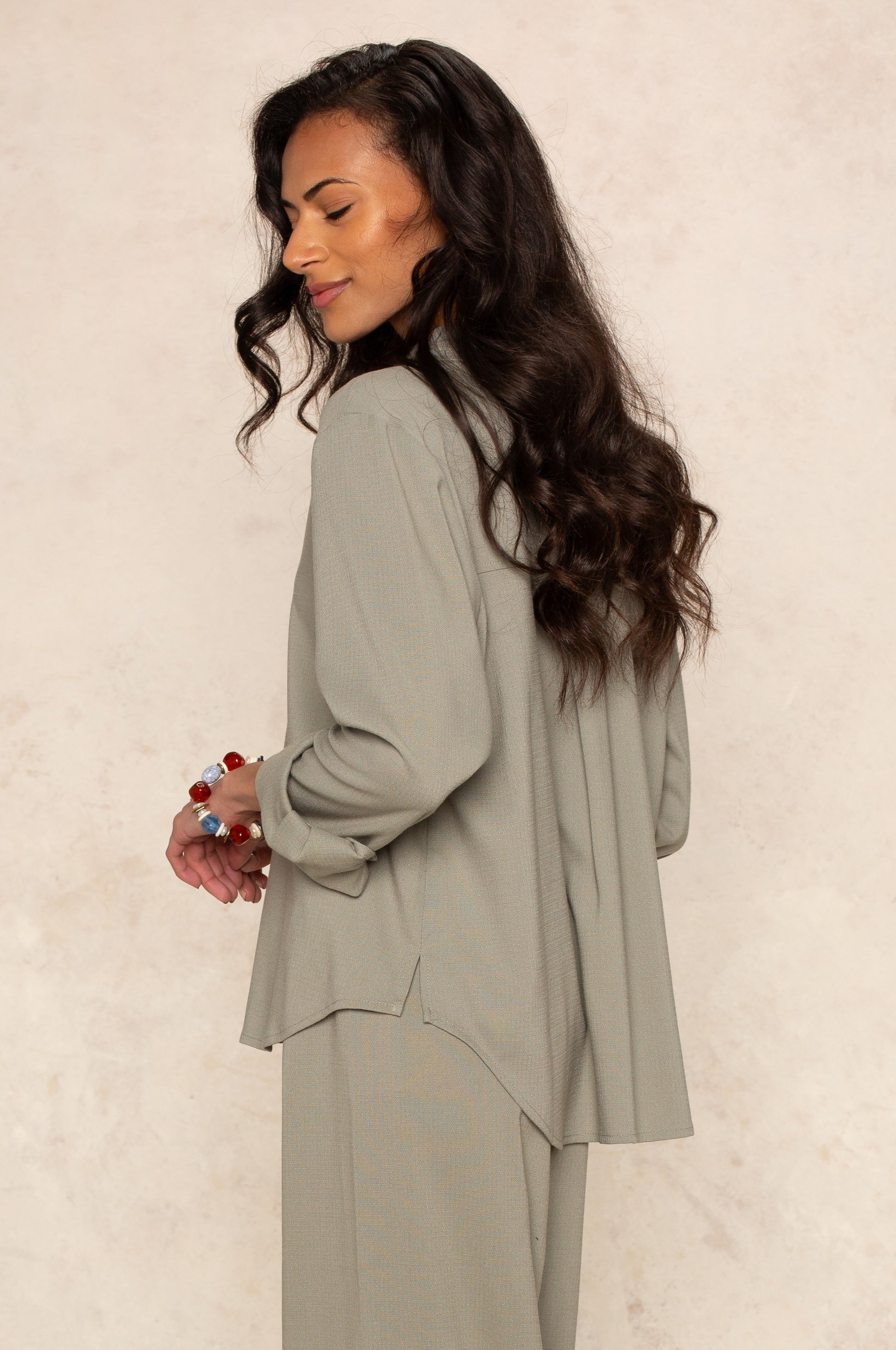 A woman with long, wavy dark hair smiles with her eyes closed, wearing the Georgie Seaside Blouse—loose-fitting and light gray—paired with colorful rings, posed against a softly textured background.