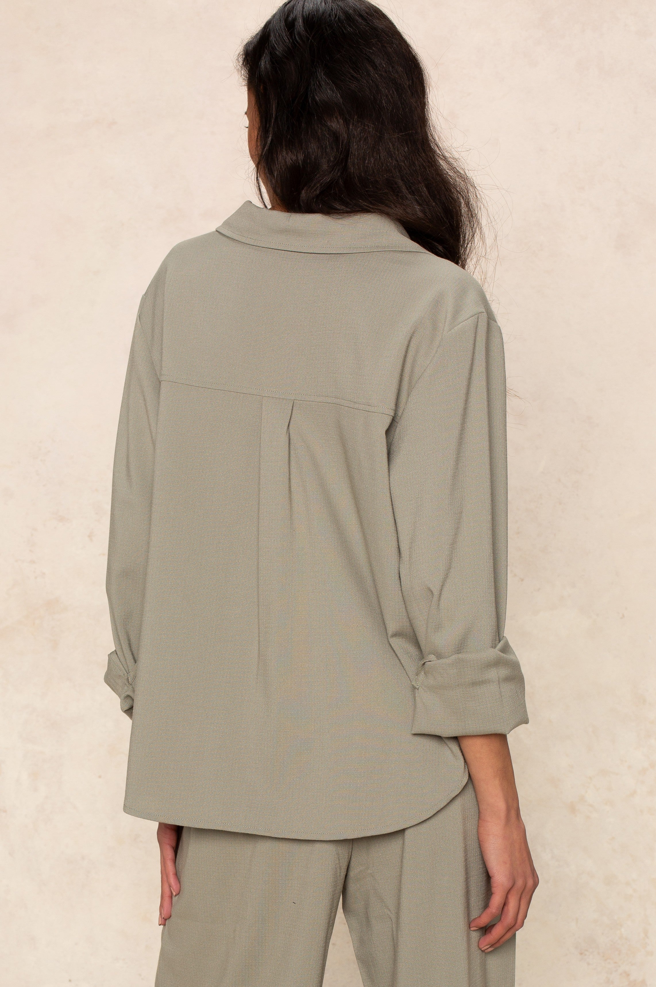 A person with long dark hair, dressed in Georgie's Seaside Blouse—loose-fitting and light gray with rolled-up sleeves—and matching pants, stands facing away from the camera against a light beige background.