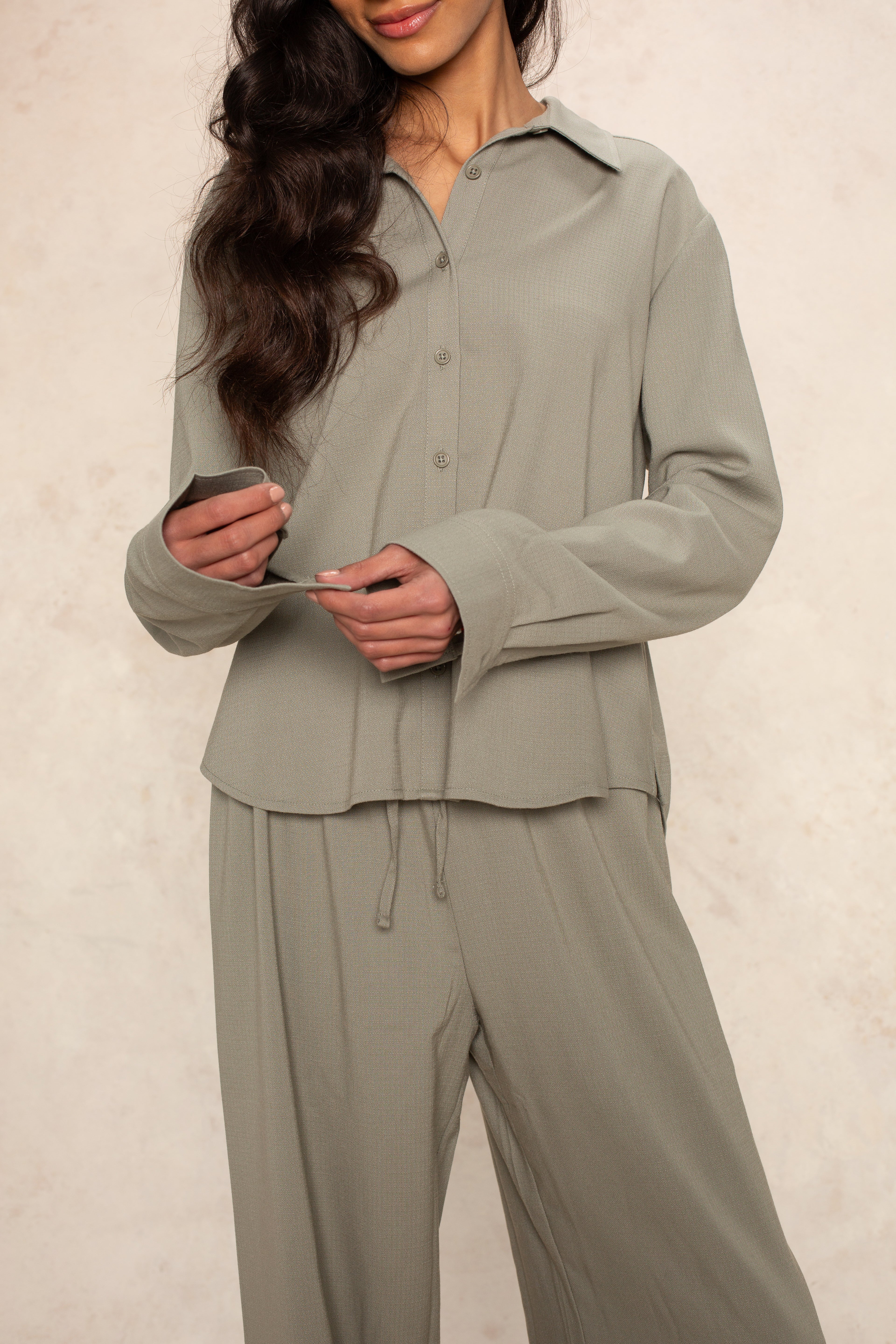 A woman models the Georgie Seaside Blouse, a loose-fitting, light gray button-up shirt with matching wide-leg pants, standing against a neutral backdrop. Her long wavy hair drapes over one shoulder, face partially out of frame.