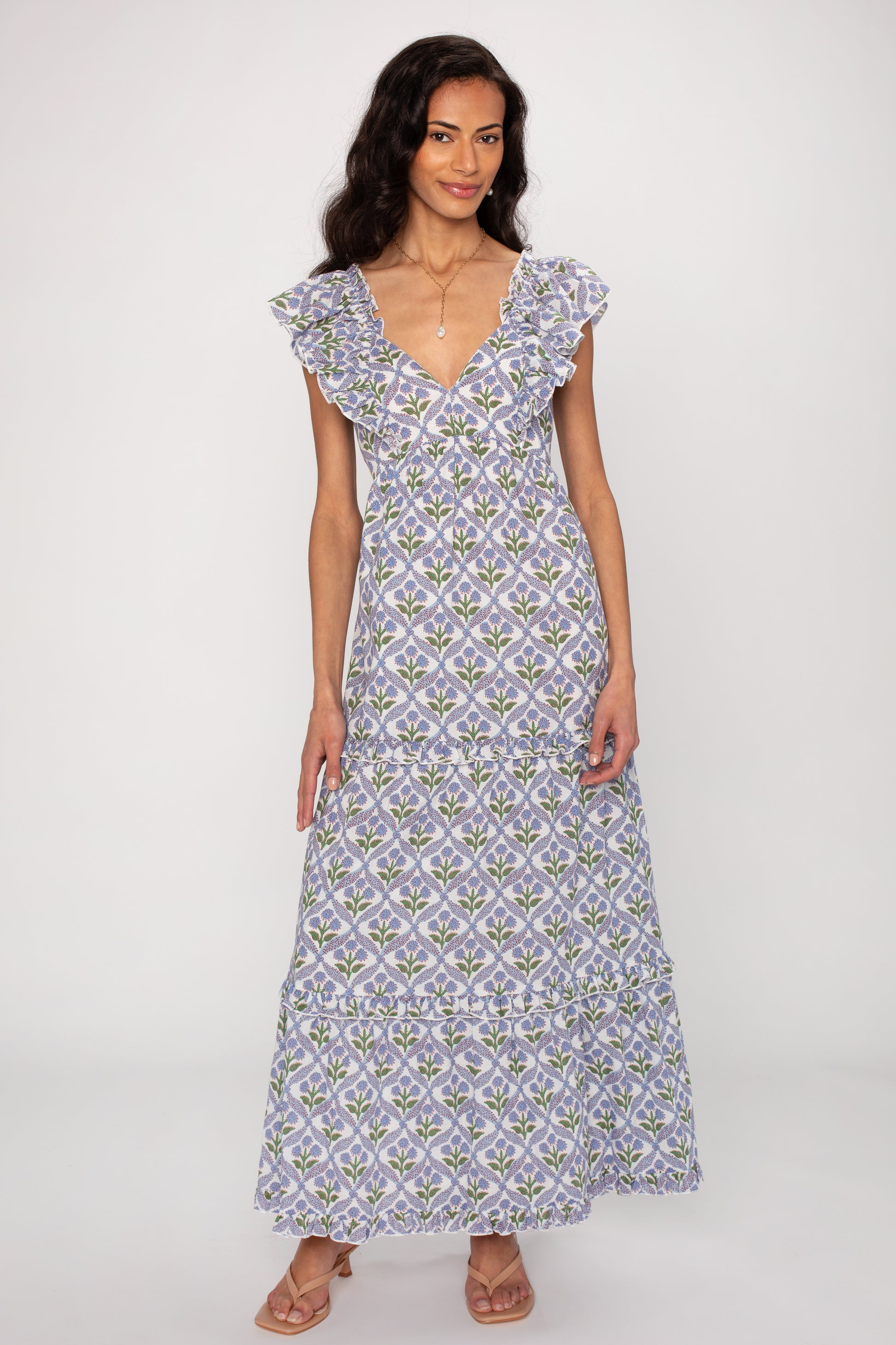A woman models the Saylor Carolina Maxi Dress, a sleeveless, floor-length style with a purple, green, and white floral print and ruffled V-neckline. Its flowing silhouette and lightweight fabric make it a versatile wardrobe essential.