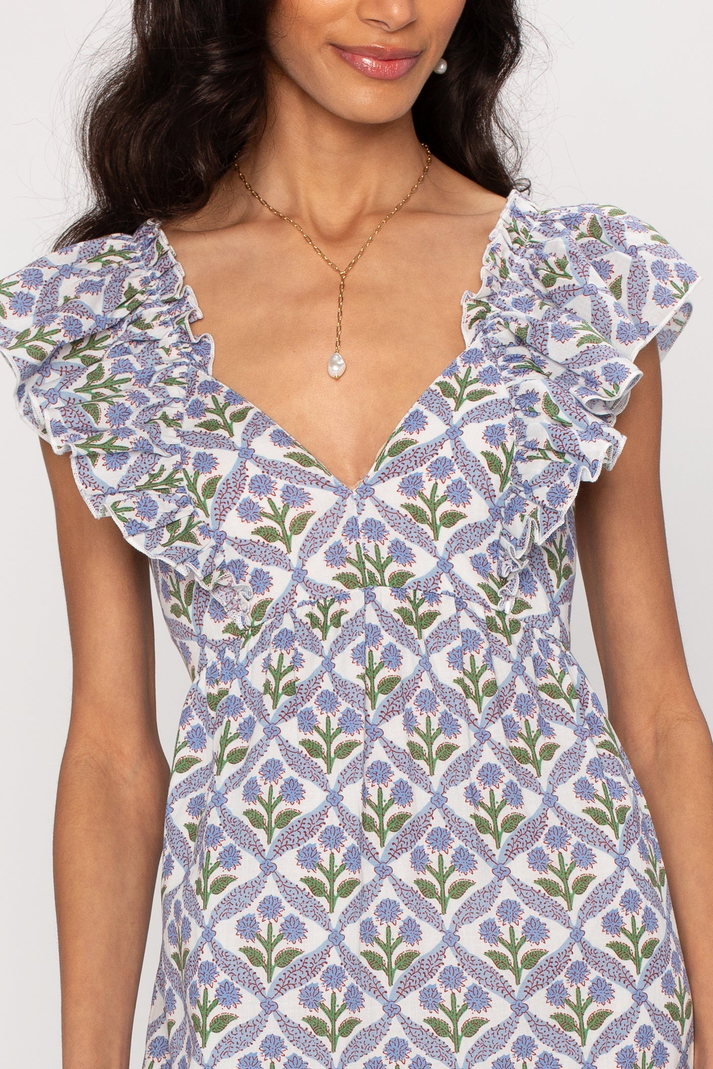 A woman wears the Saylor Carolina Maxi Dress, a v-neck style with ruffled sleeves and a blue, green, and purple floral print in lightweight fabric for a flowing silhouette. She pairs it with a gold chain necklace with a teardrop pendant.