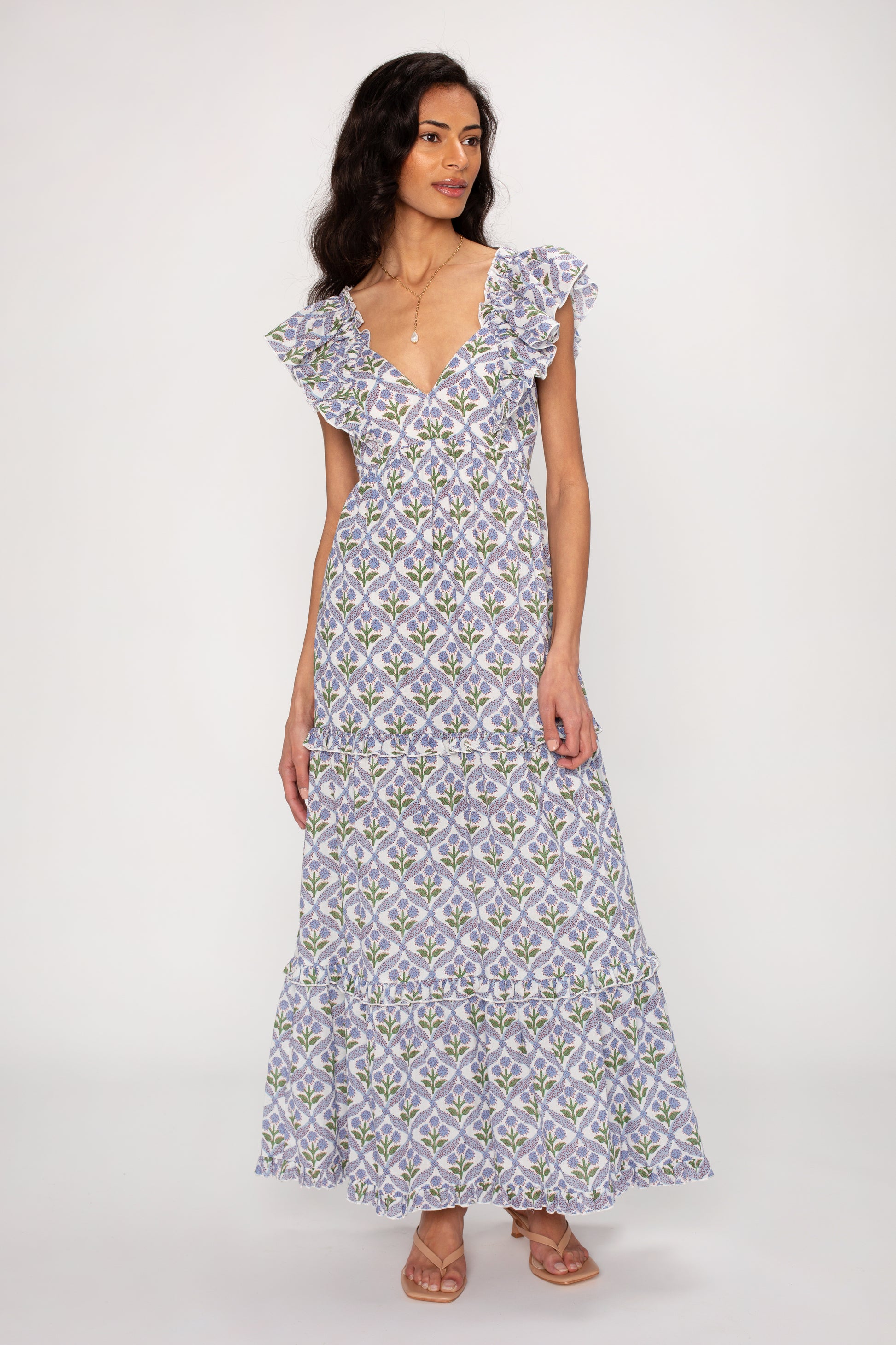 A woman wears the Carolina Maxi Dress by Saylor—a sleeveless, floor-length dress in purple and green florals with ruffled sleeves and a tiered skirt. Made from lightweight fabric, its flowing silhouette completes her look as she smiles gently.