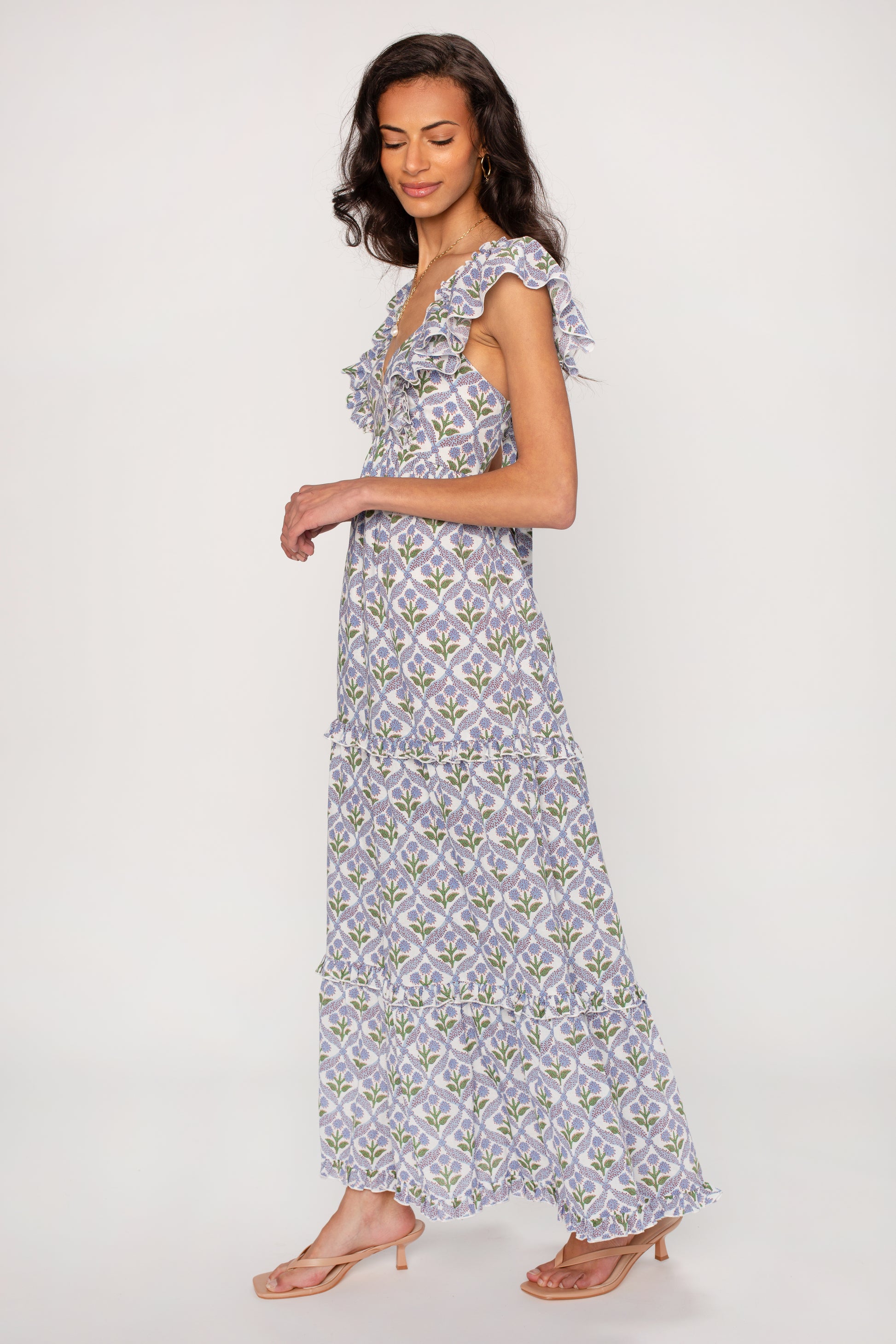 A woman stands in profile wearing the Saylor Carolina Maxi Dress, a long sleeveless summer dress with a flowing silhouette and ruffled details. Her wavy hair frames a plain, light backdrop as she pairs the versatile piece with sandals.