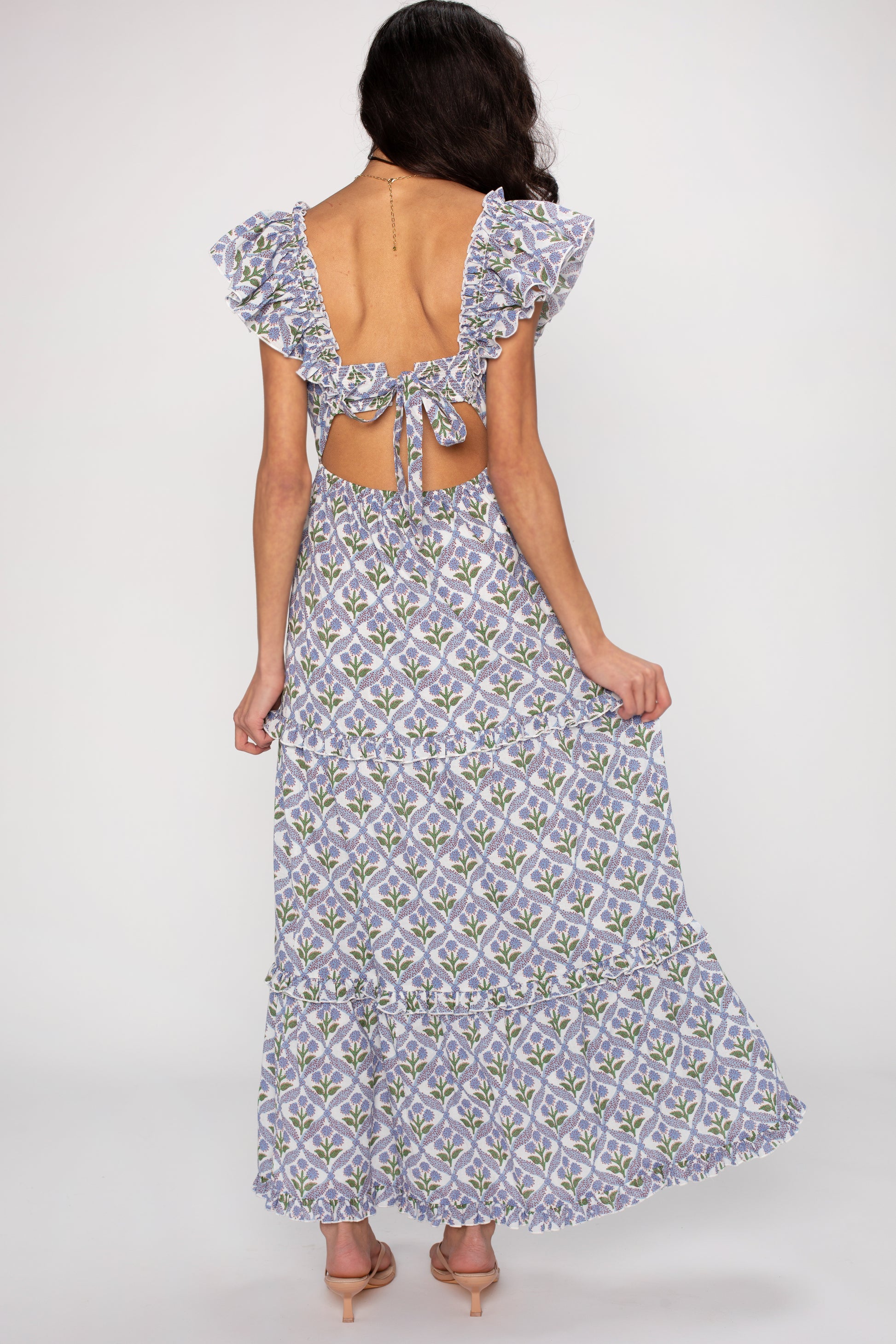 A woman faces away, wearing the Saylor Carolina Maxi Dress—a light-hued, floral dress with ruffle sleeves, open back, and bow tie detail. Its flowing silhouette complements her wavy dark hair and nude heels for a versatile look.