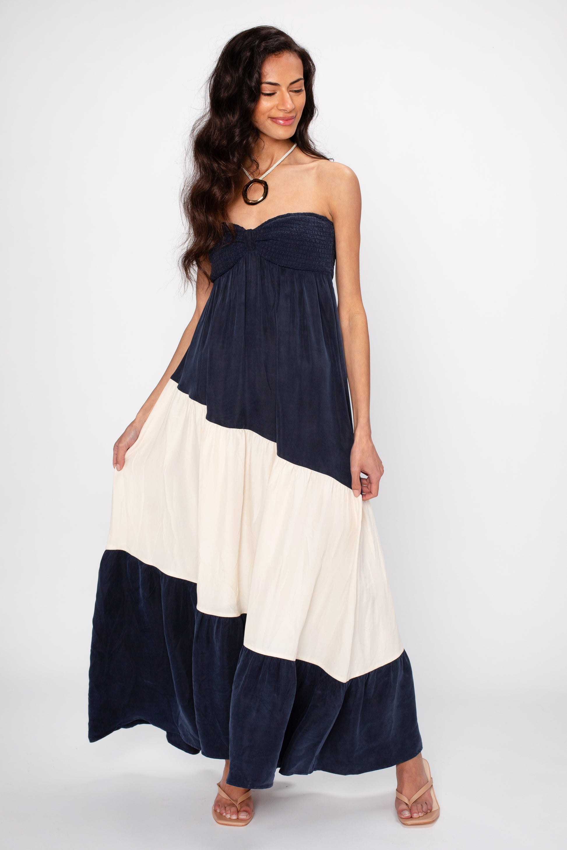 A woman with long dark hair wears The Sashia Maxi Dress by Sancia— a strapless, navy and white color-block, maxi-length dress— with nude heels. She stands against a white background, smiling gently and holding the sides of her dress.