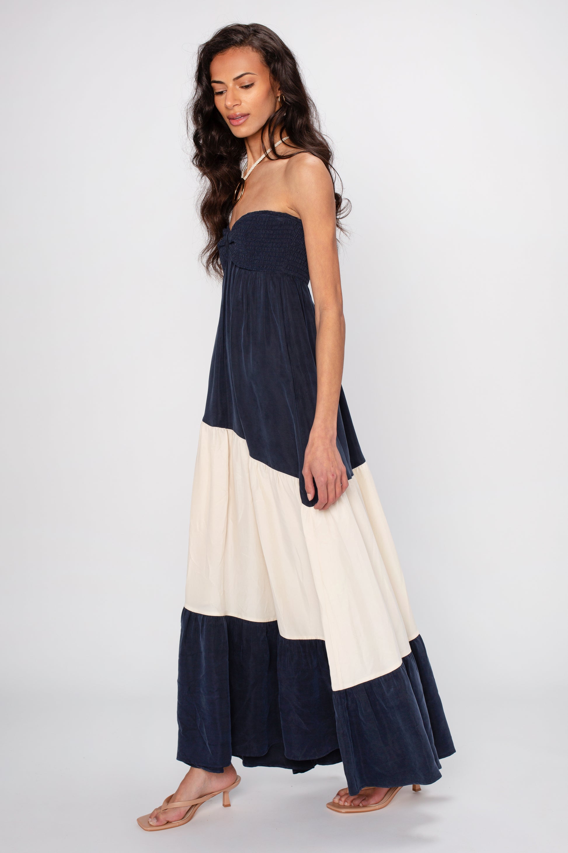 A woman wearing the Sashia Maxi Dress by Sancia—strapless, navy blue and white color-blocked, and maxi length—stands sideways with long wavy hair, nude heeled sandals, and a plain white background.