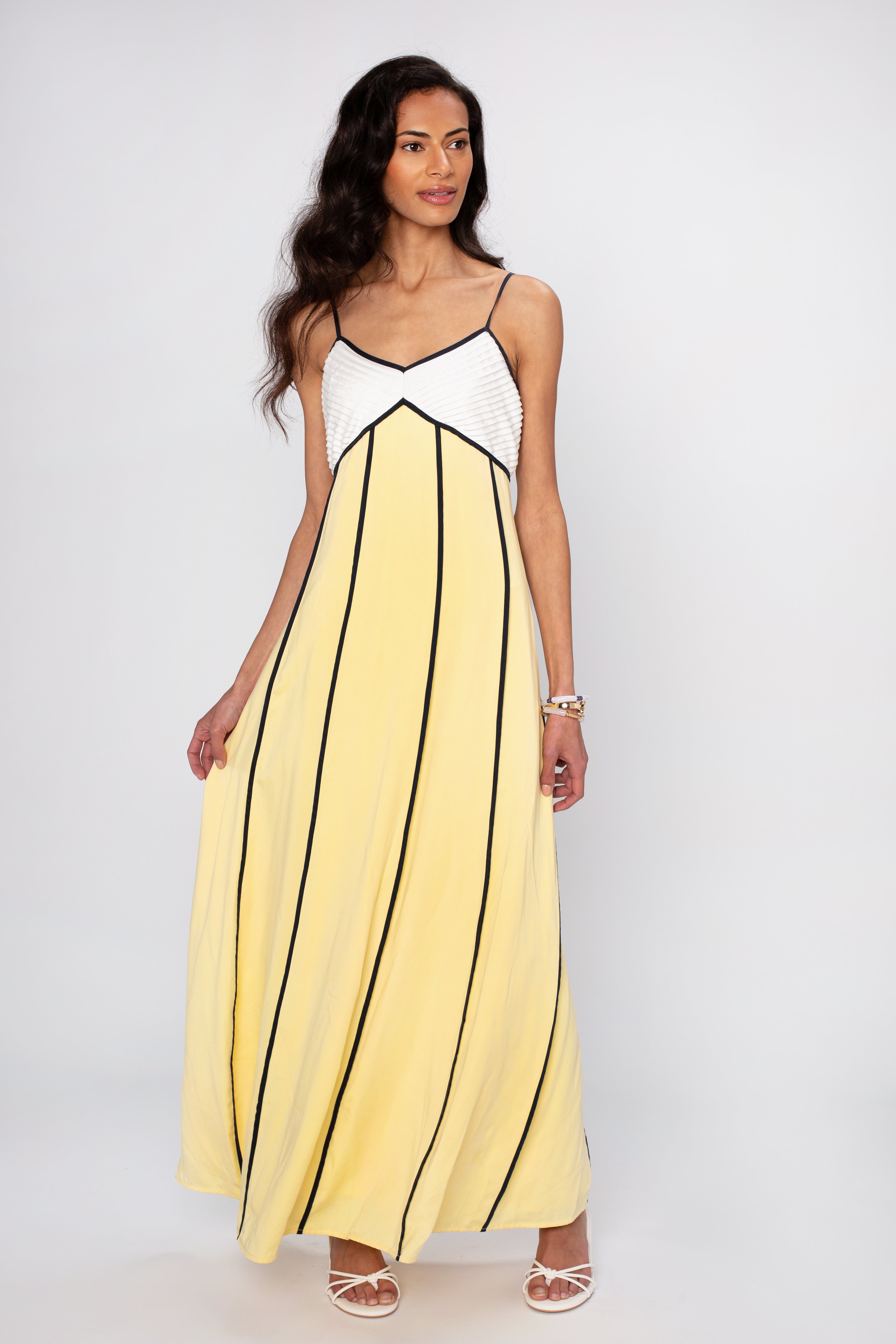 A woman stands against a plain light background, holding the side of The Odele Maxi Dress by Sancia—a relaxed fit, sleeveless yellow maxi dress featuring black vertical lines and a chic white top detail.