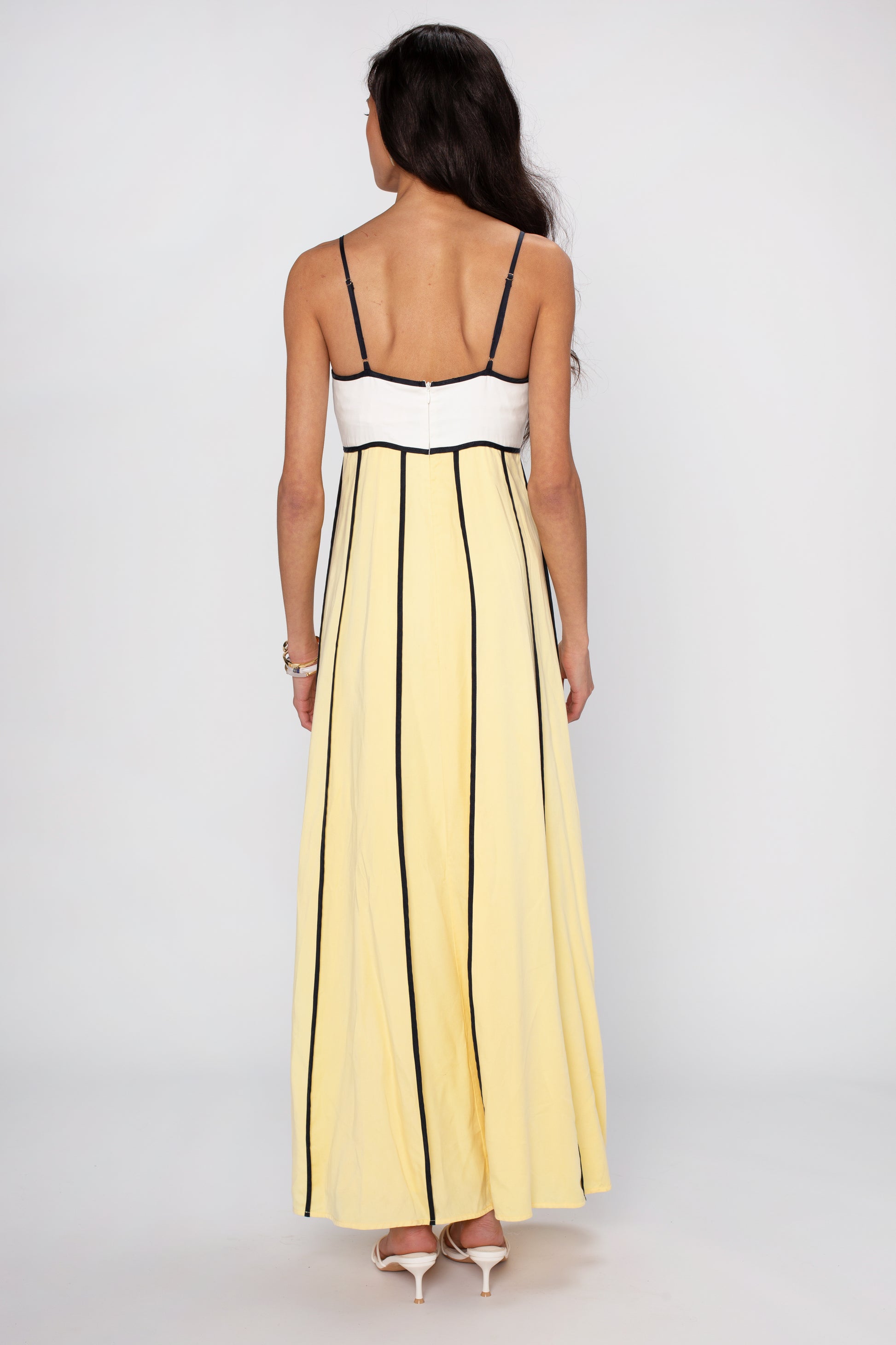 A woman with long dark hair wears The Odele Maxi Dress by Sancia, featuring a sleeveless relaxed fit, yellow and black vertical stripes, and a white top section. She pairs it with white heels against a plain light background.