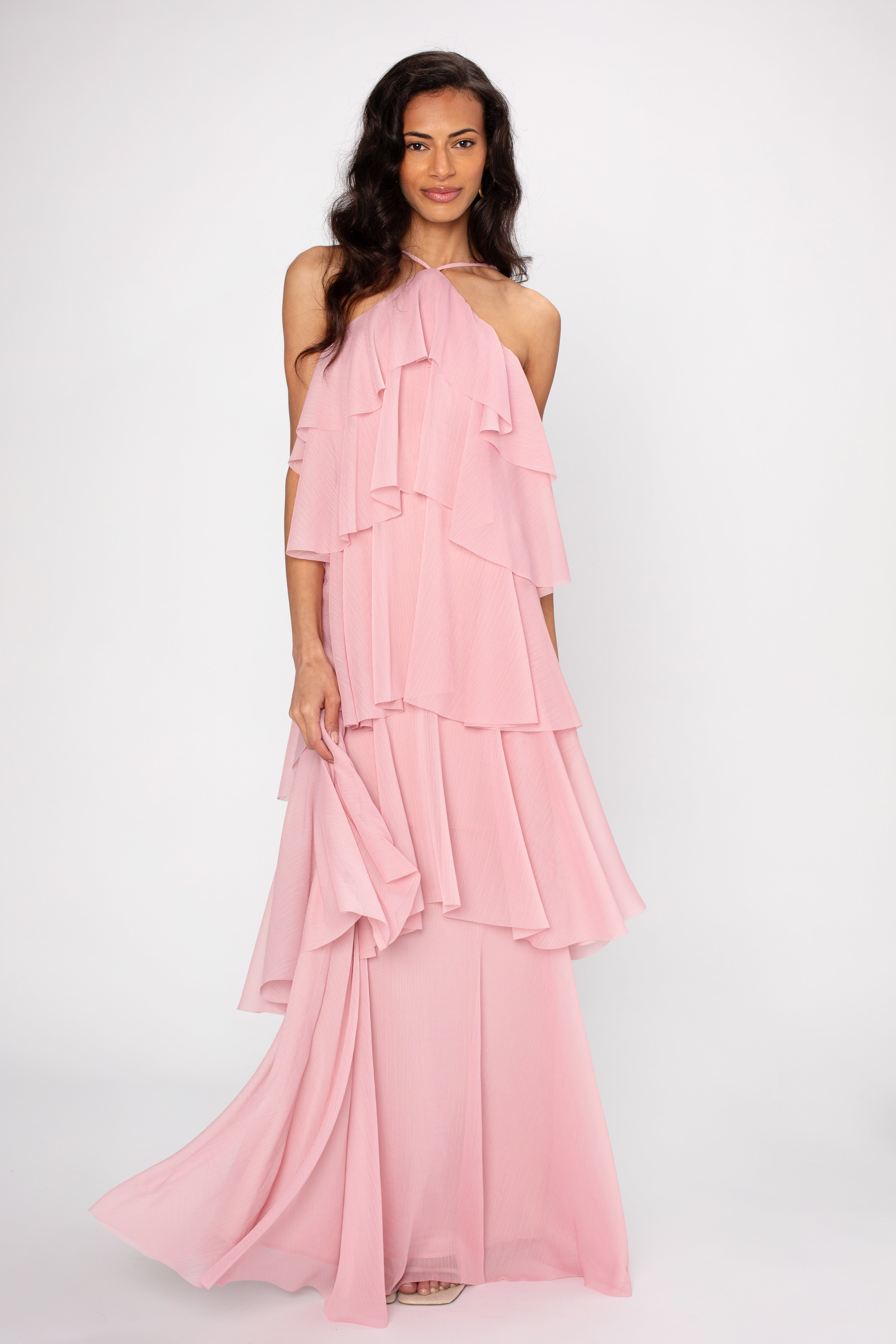A woman with long, wavy dark hair smiles gently at the camera while wearing the Georgie Ophelia Ruffle Tiered Maxi Dress in pink, standing against a plain white background.