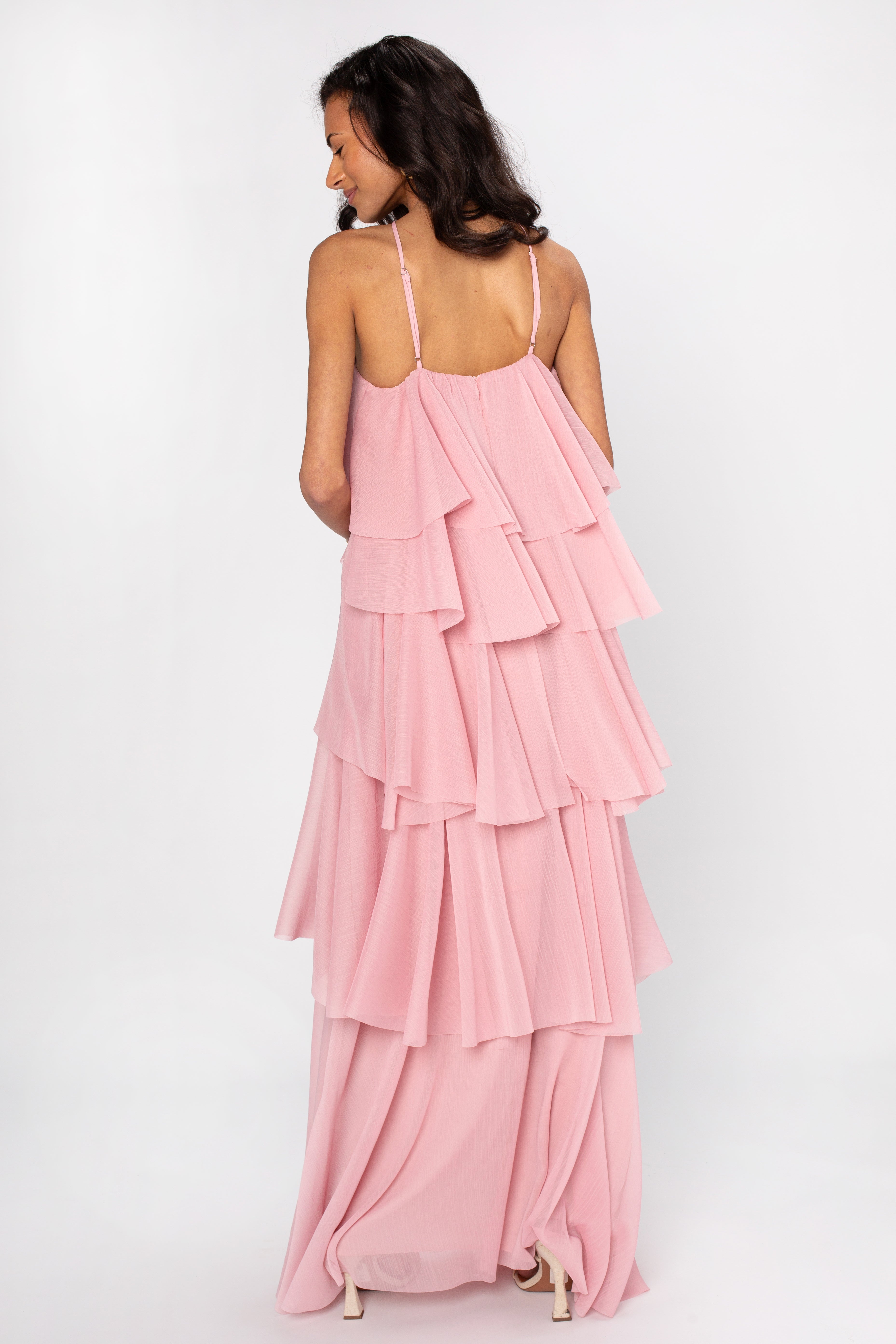 A woman faces away in the Georgie Ophelia Ruffle Tiered Maxi Dress, a flowing pink maxi with thin straps and cascading ruffles, set against a plain white background—an elegant choice for evening wear.