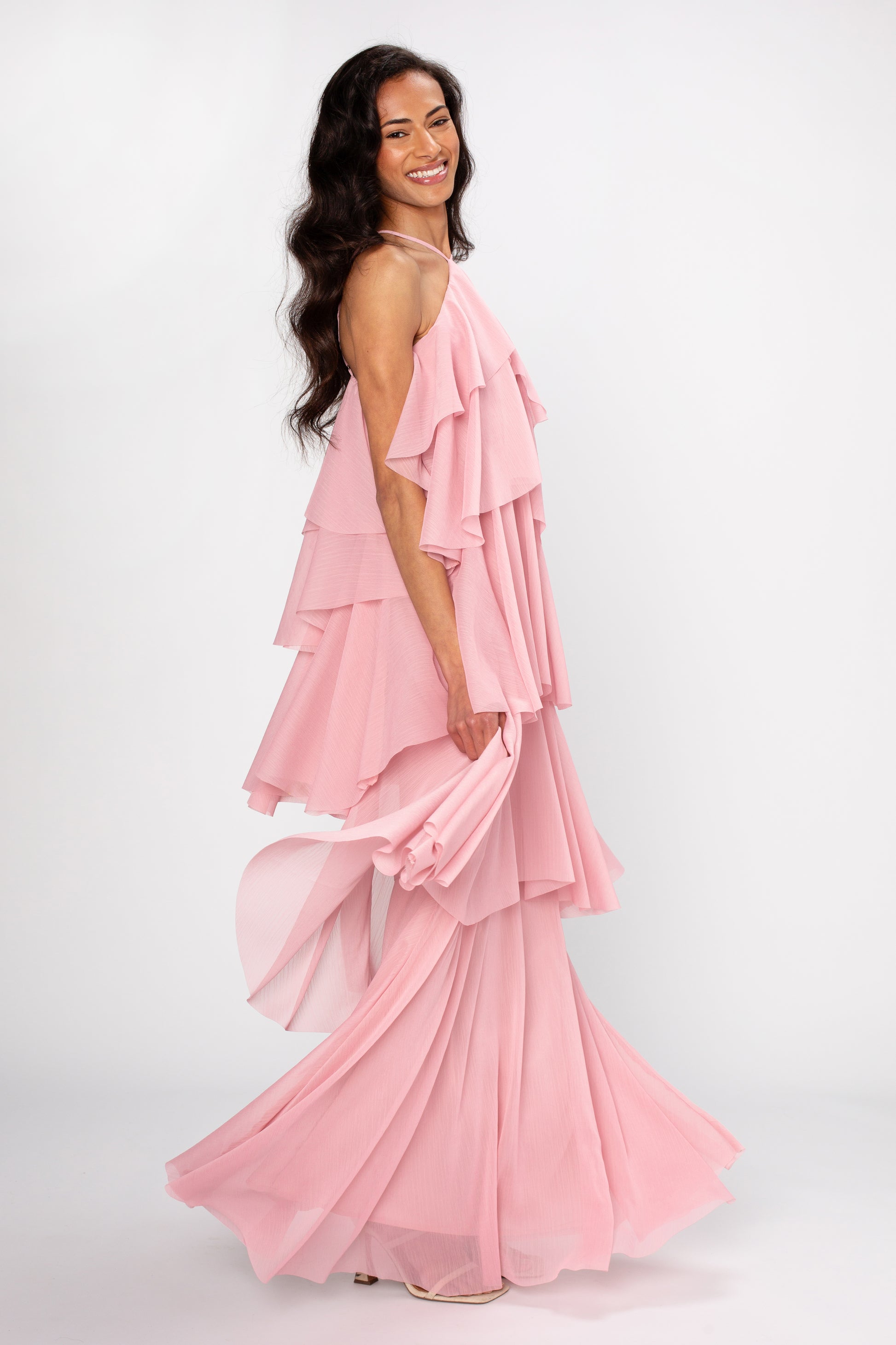 A woman with long dark hair smiles while wearing the Georgie Ophelia Ruffle Tiered Maxi Dress in pink and light heels, standing against a plain white background.