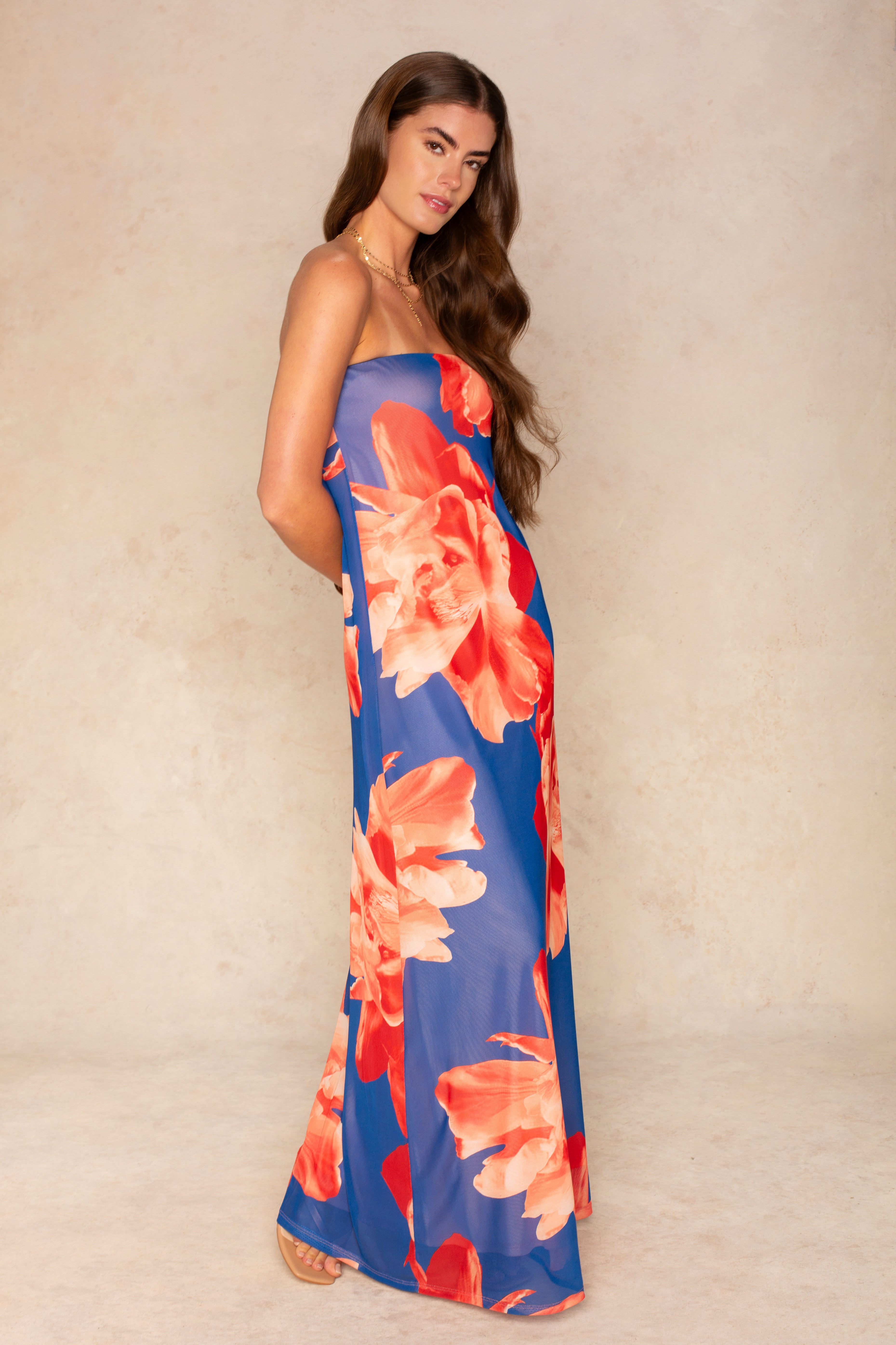 A woman with long brown hair models the Isla Flora Tube Mesh Maxi Dress, featuring bold red and orange floral prints on blue—perfect for summer evenings—while standing sideways against a beige background.